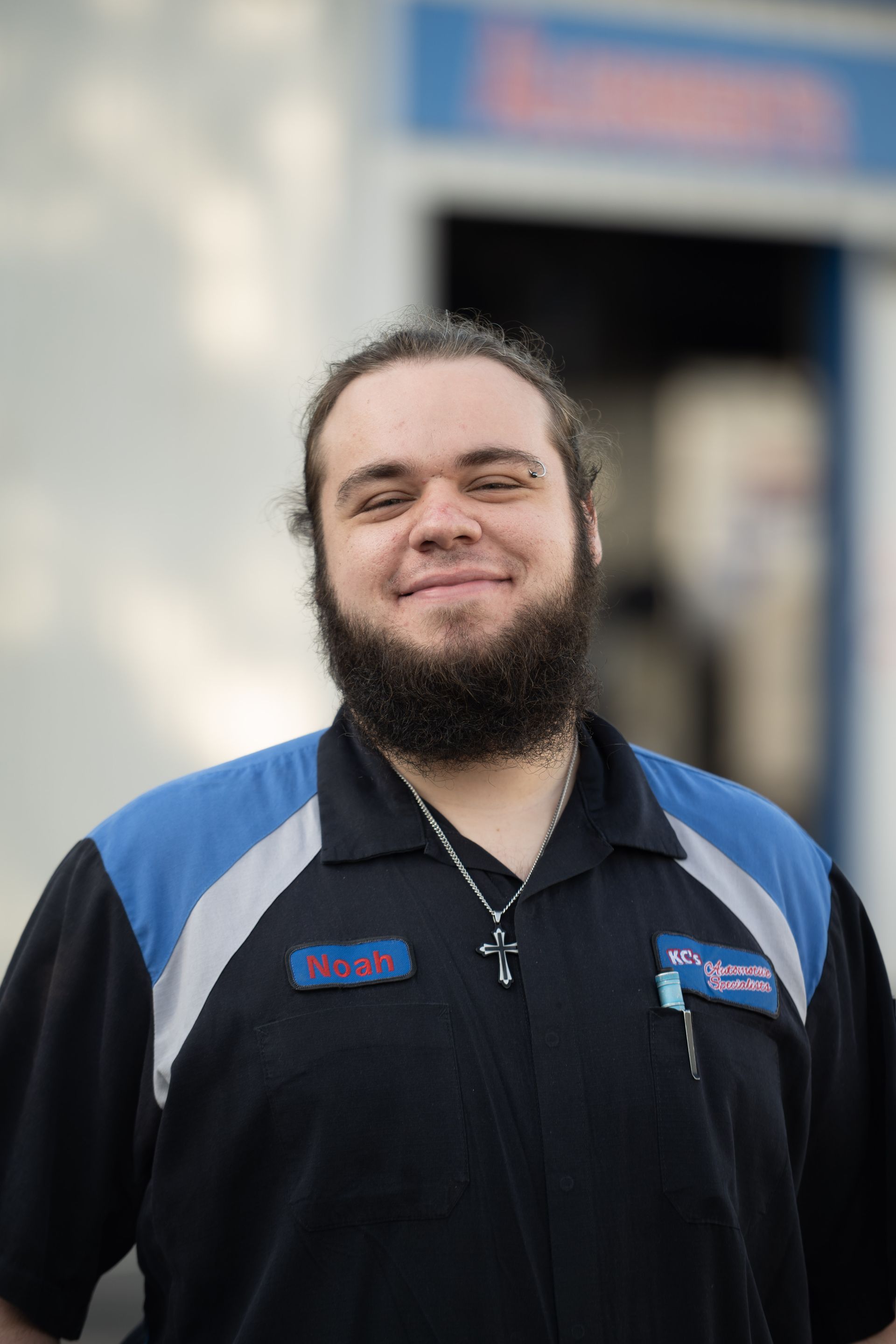 Mechanic in blue and black uniform smiles, standing outside a garage.