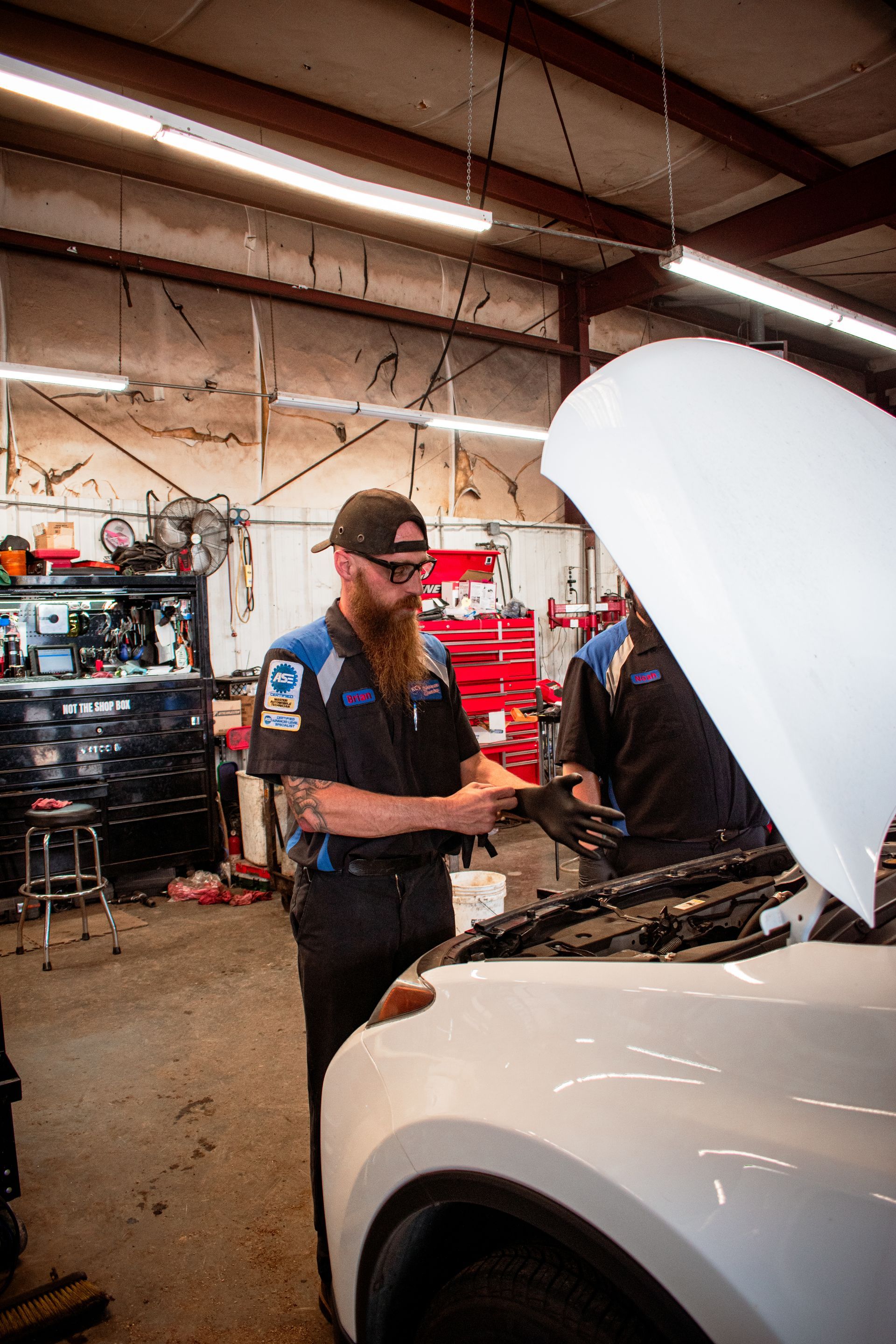 Mechanic in blue uniform inspects engine, another visible, in a repair shop with tools and a raised hood on a white car.
