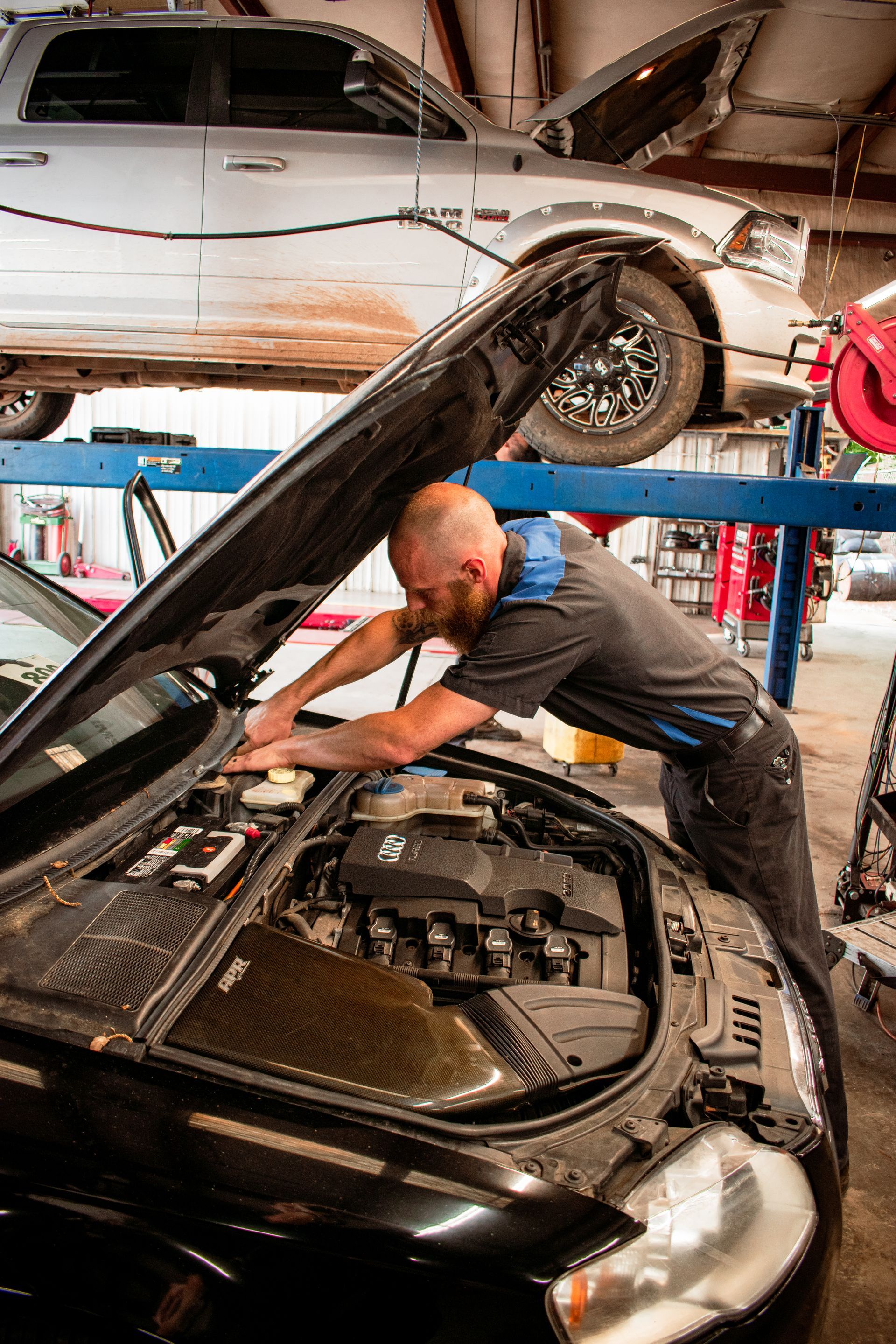 Mechanic works on a black car with the hood open in a garage. Another car is on a lift in the background.