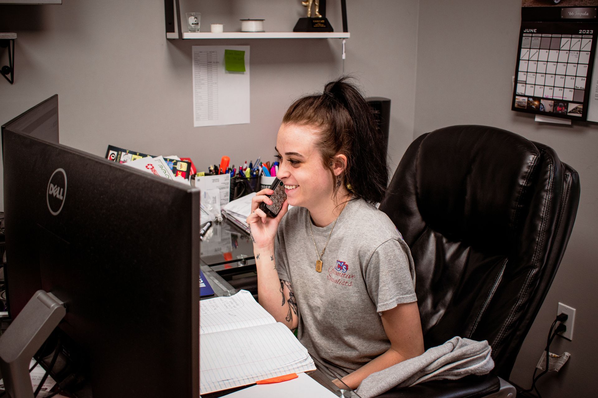 Woman smiling while talking on a phone at a desk with computer and paperwork in an office setting.
