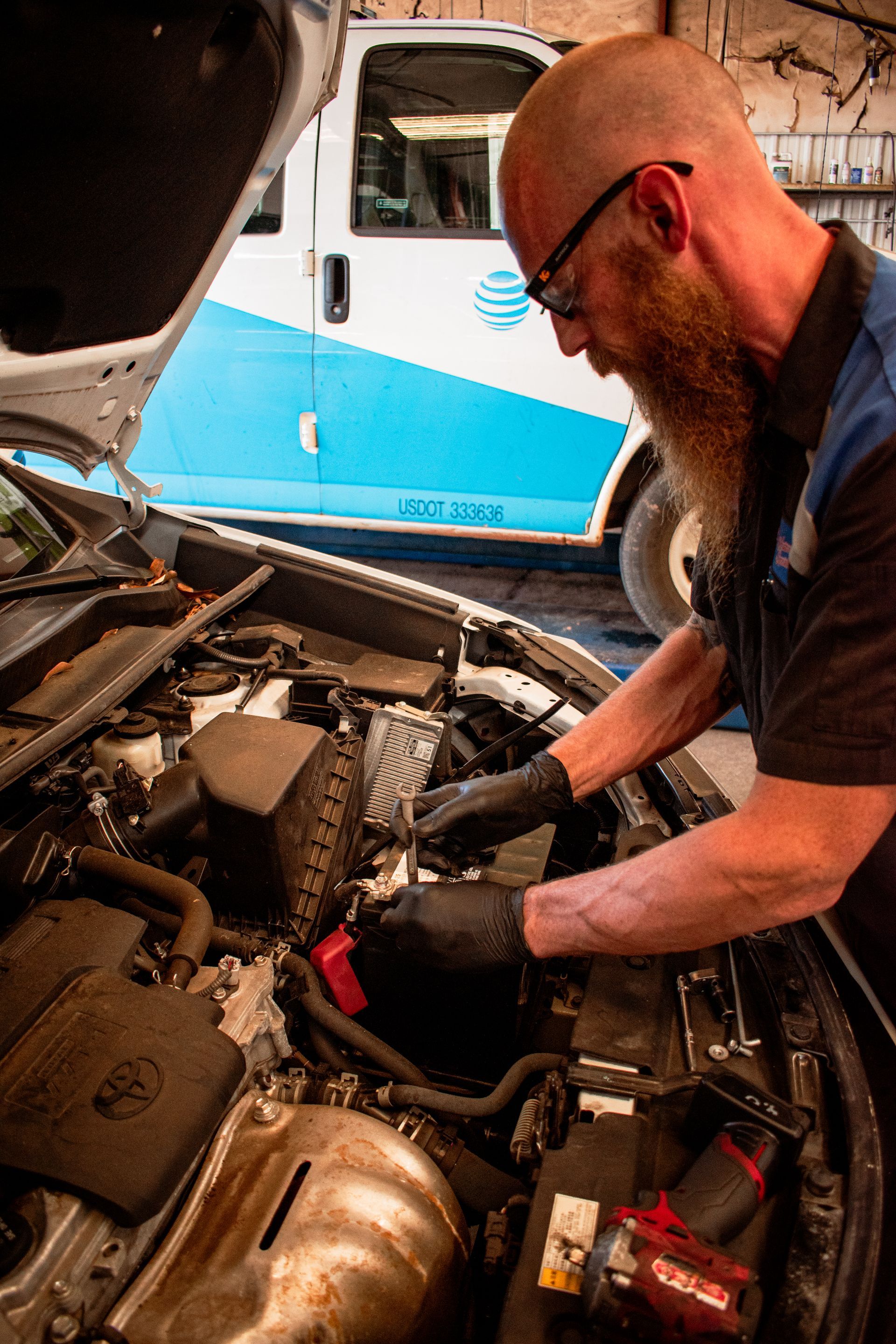Mechanic works on a car engine; a white AT&T van is in the background.