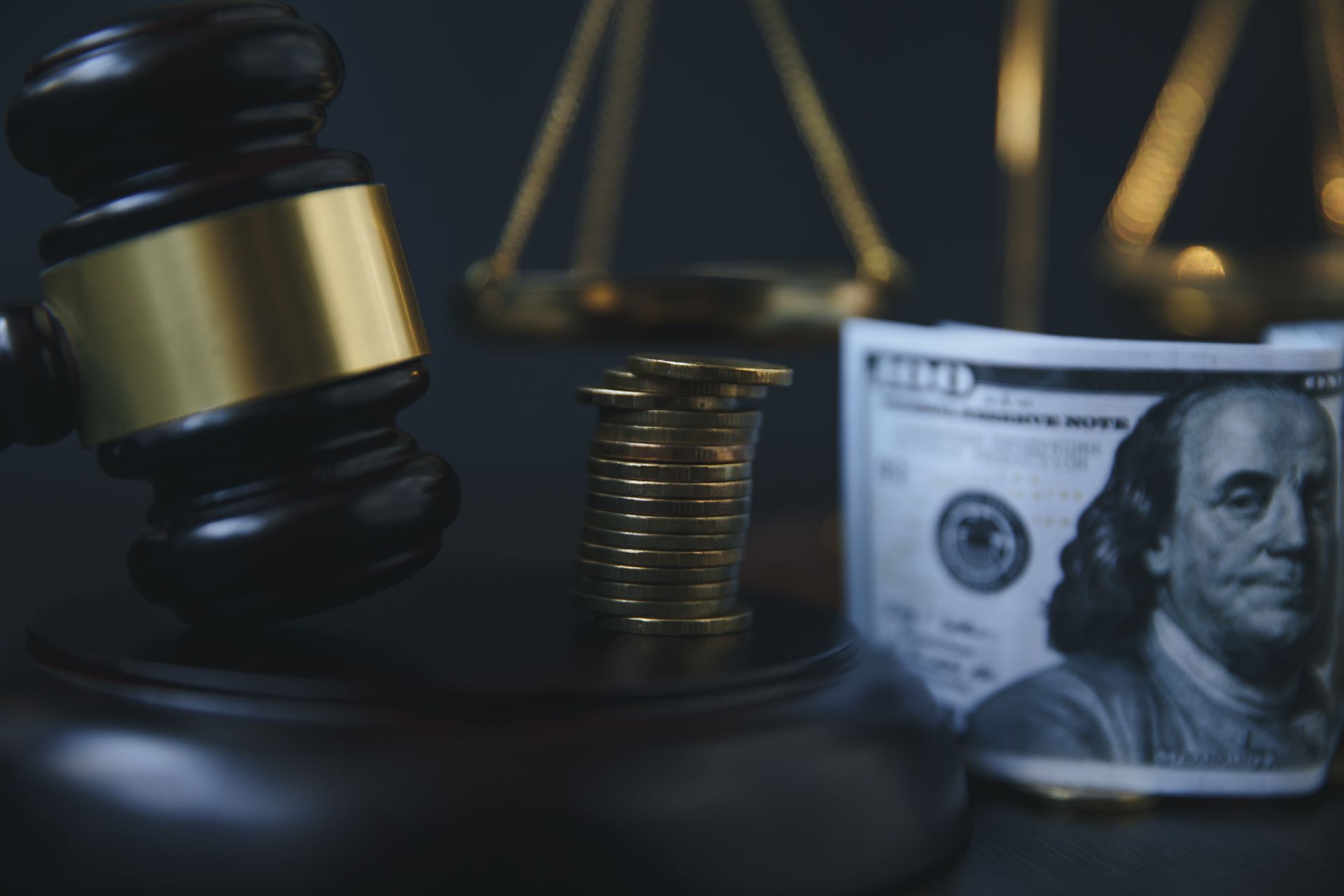Gavel, scales of justice, US currency, and stack of coins on dark surface, symbolizing legal and financial matters.