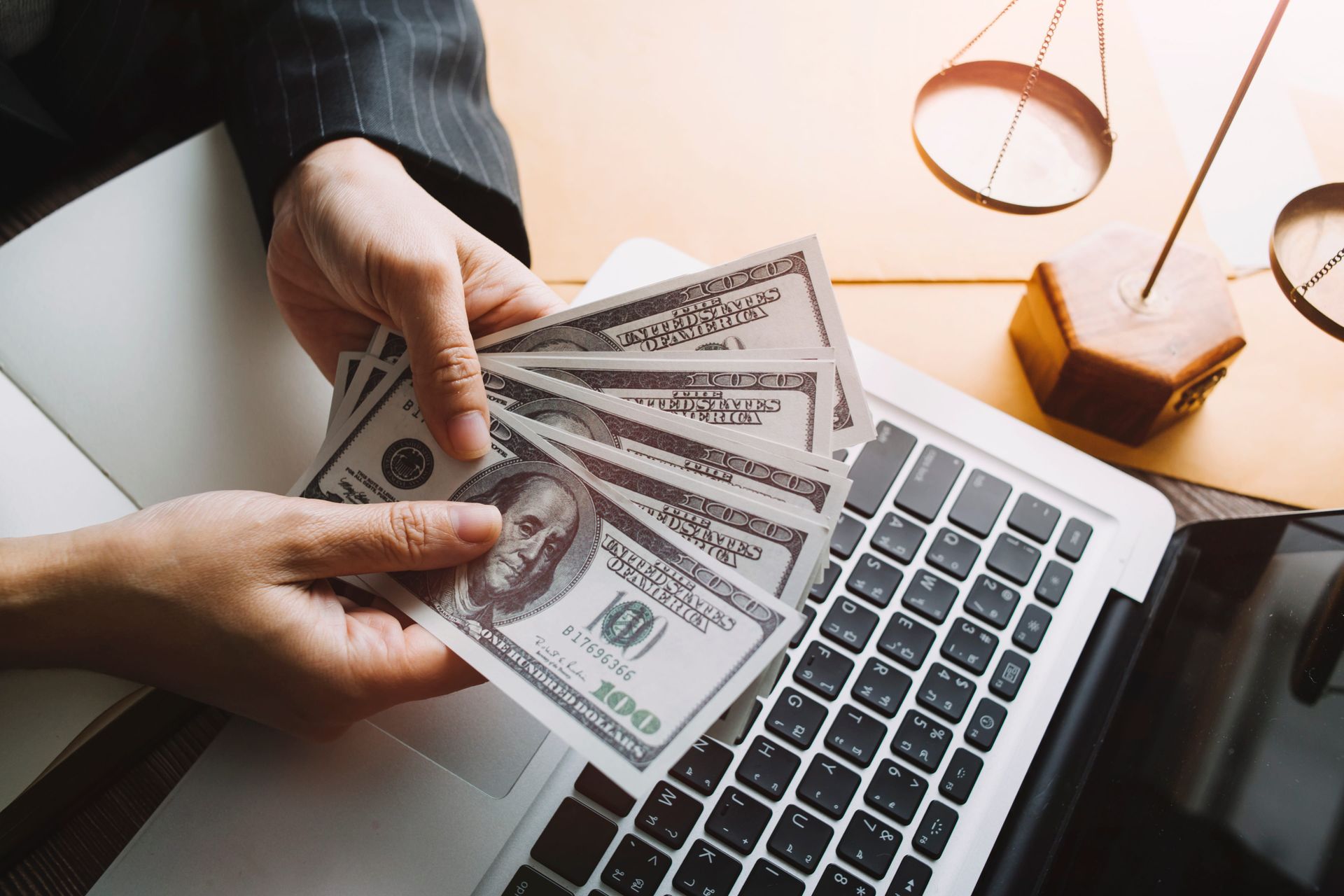 Hands counting U.S. dollar bills over a laptop keyboard with coffee and notebook nearby