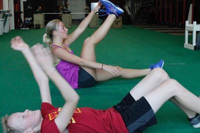 Two people stretching on green gym mats in a gym in Urbandale, IA, one holding a blue shoe overhead, the other lying on their back.