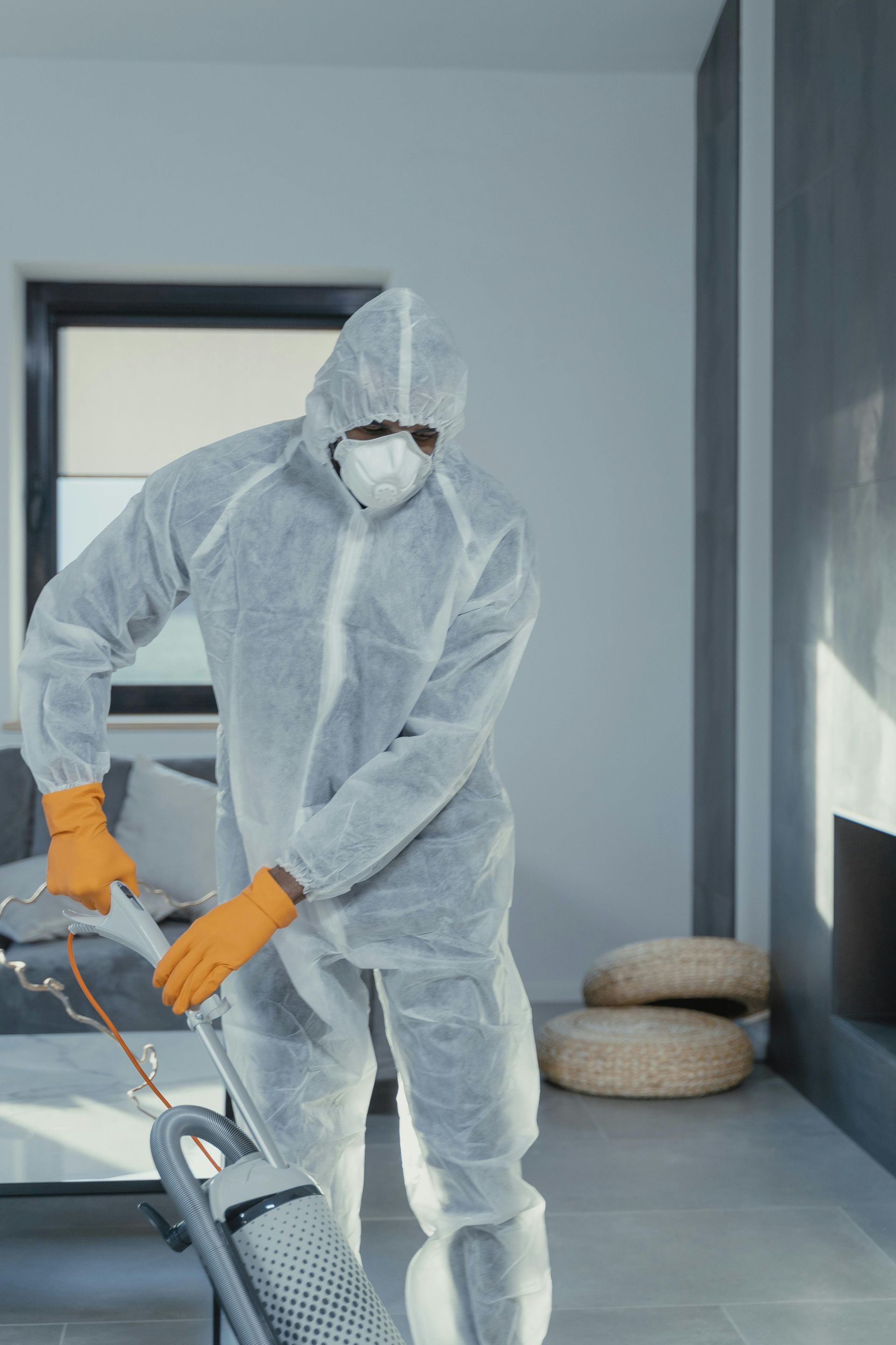 Person in hazmat suit vacuuming a room. They wear orange gloves and a white mask.