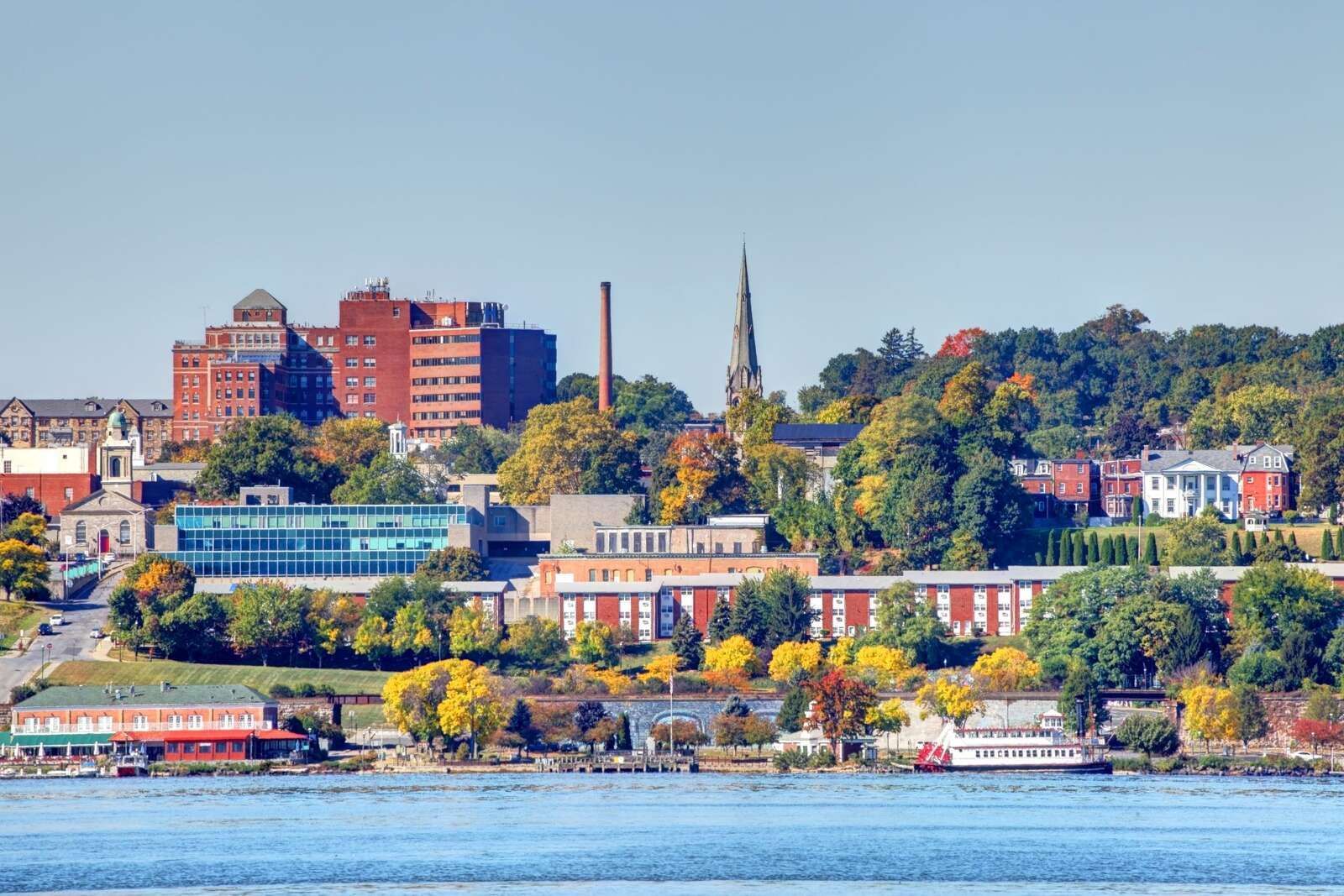 Waterfront city skyline with brick buildings, church spires, and autumn trees under a clear blue sky