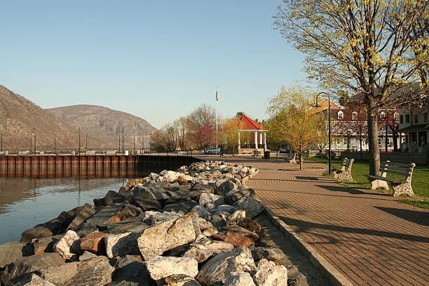 Riverfront promenade with rocks, benches, trees, and calm water beside mountains at sunset