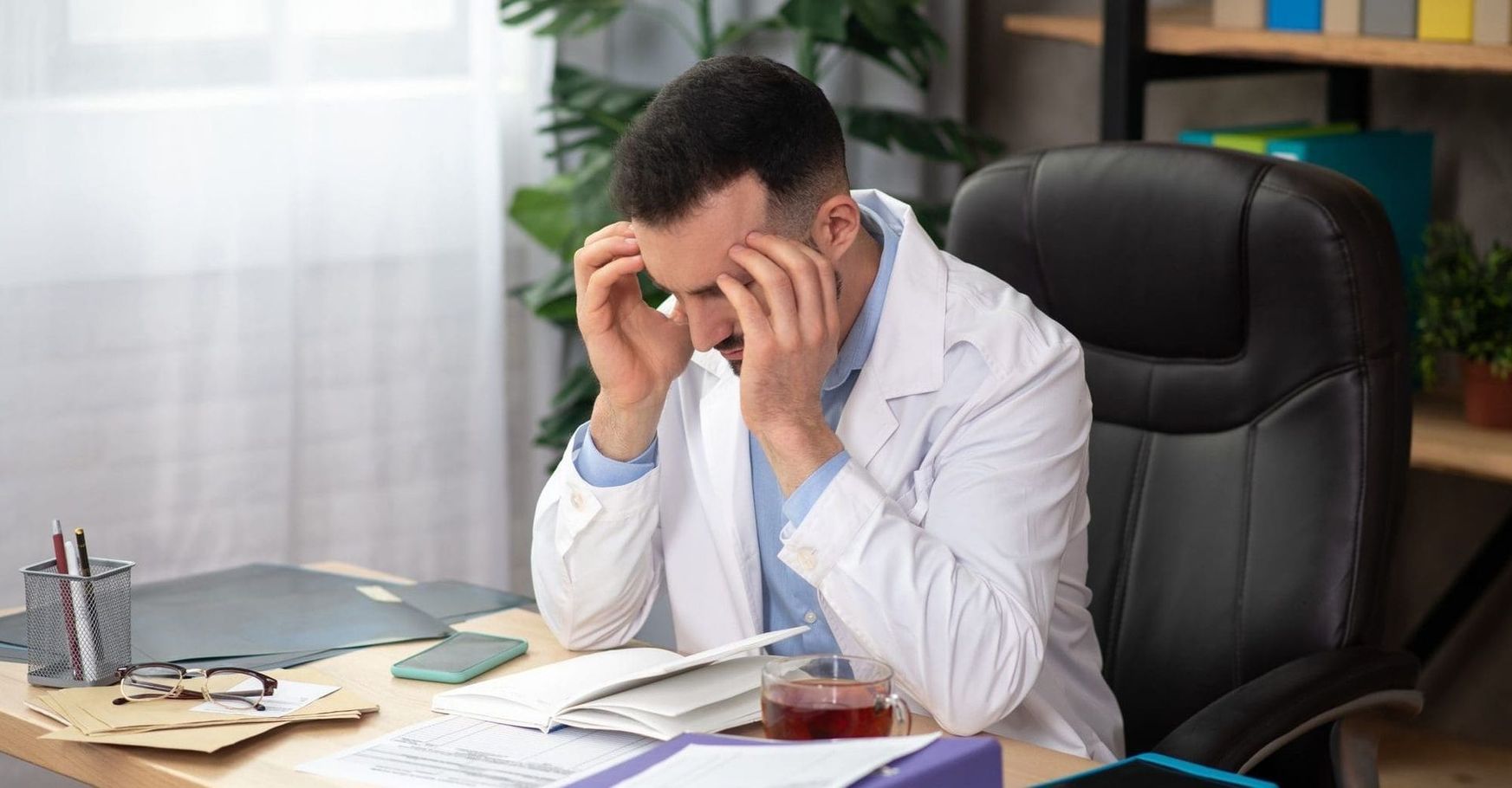 Stressed man in a white shirt sits at a desk, rubbing his temples amid scattered papers.