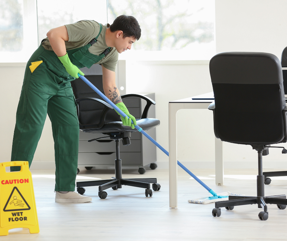 Cleaner mopping office floor near caution sign and desks, wearing green gloves and uniform