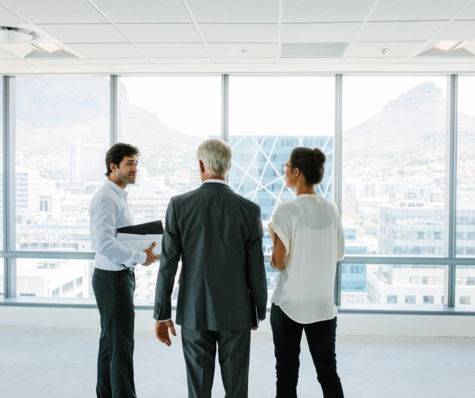 Three people stand in a bright office, looking out large windows at a city skyline.