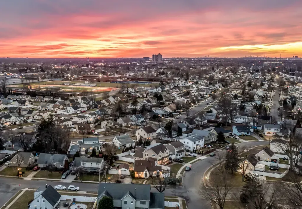 Aerial view of a suburban neighborhood at sunset with a pink-orange sky and distant city skyline