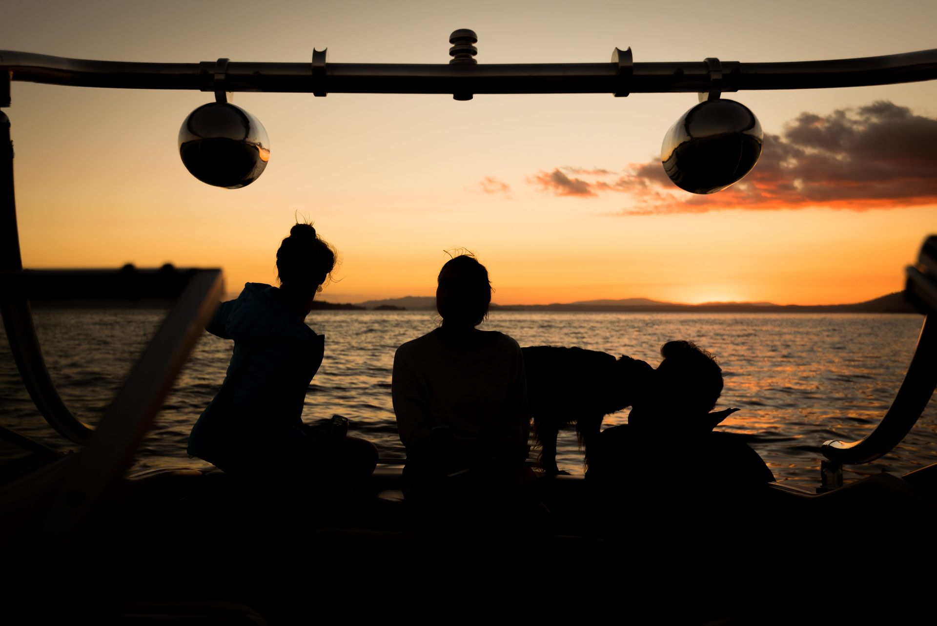 Silhouetted people sit on a boat deck at sunset, overlooking a calm, open body of water.
