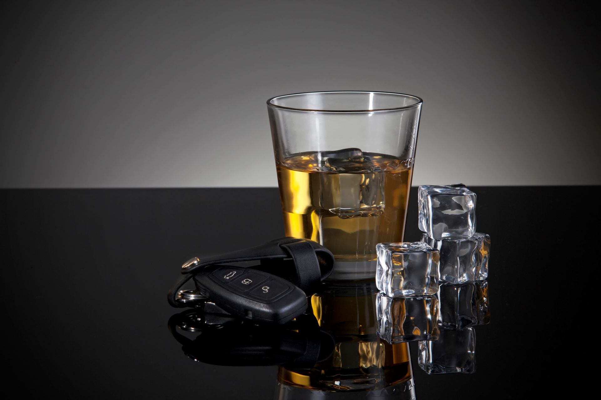 A glass of amber liquor with ice cubes sits next to a set of car keys on a reflective black surface.