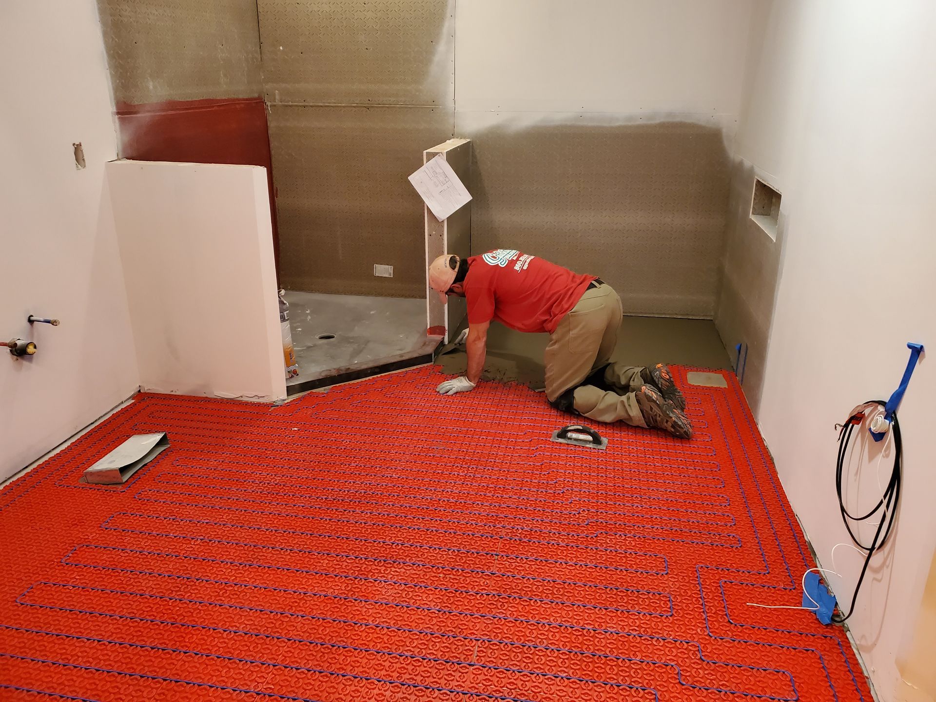 Man installing red radiant floor heating in a bathroom. Walls are white and gray, with a partially built shower.