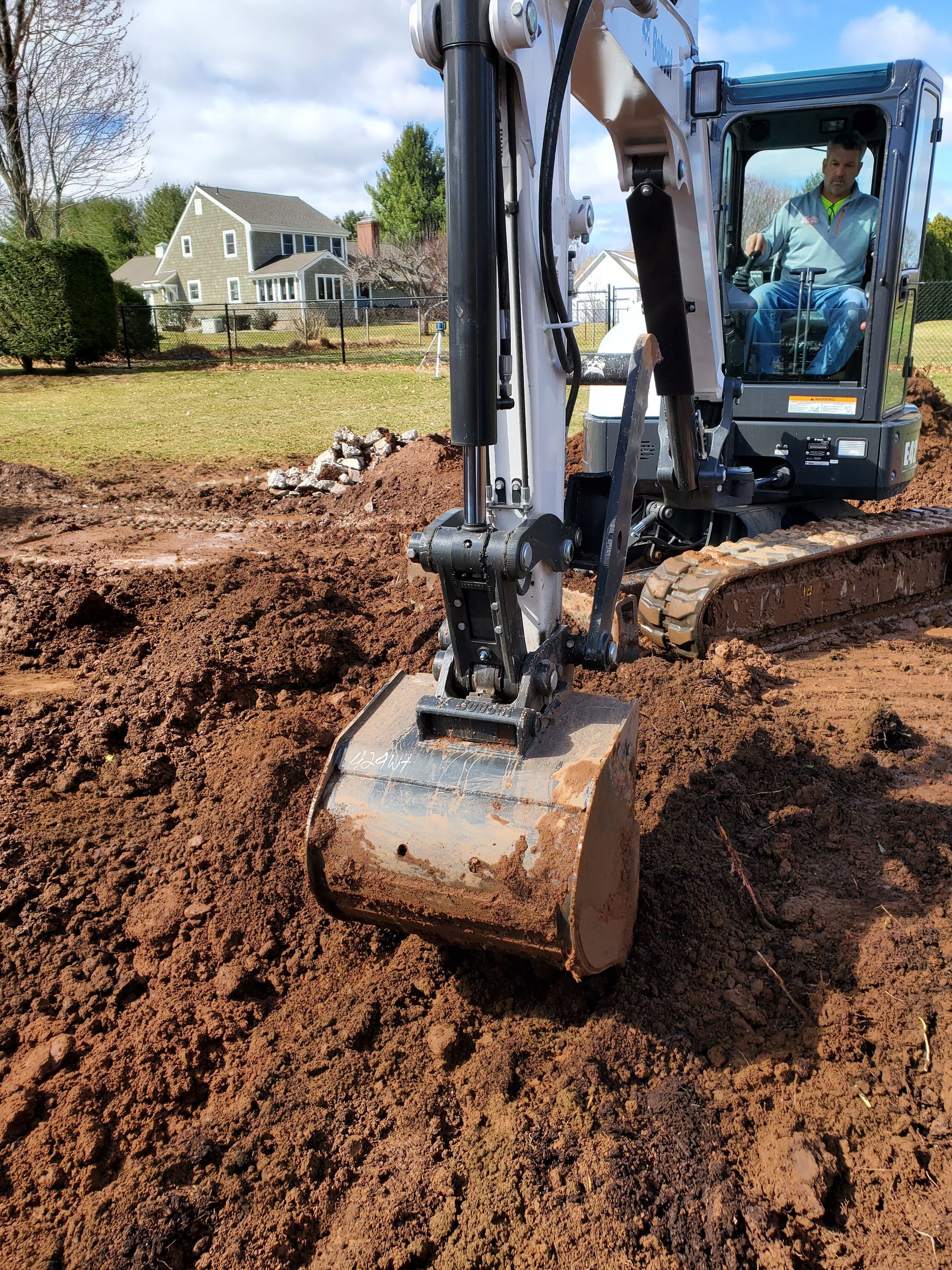 A small excavator digs in dirt; a person is inside the cab; a house is in the background.