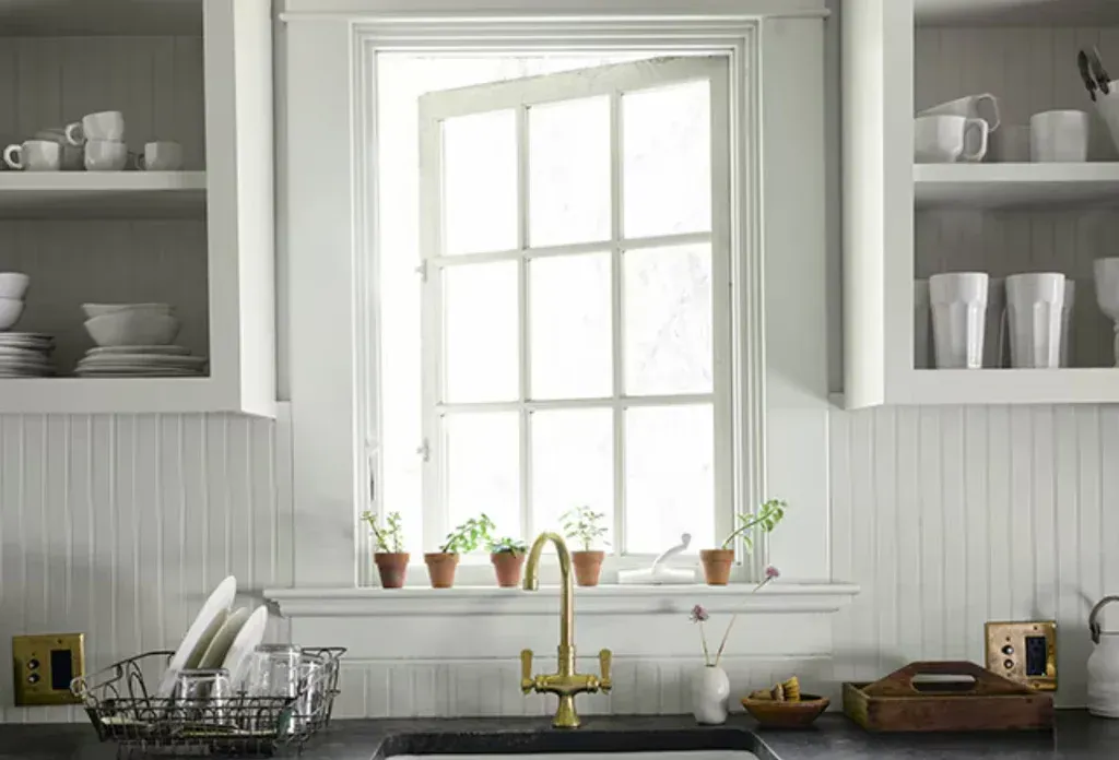 Bright kitchen window above a sink with potted herbs, white cabinets, and dishes on open shelves