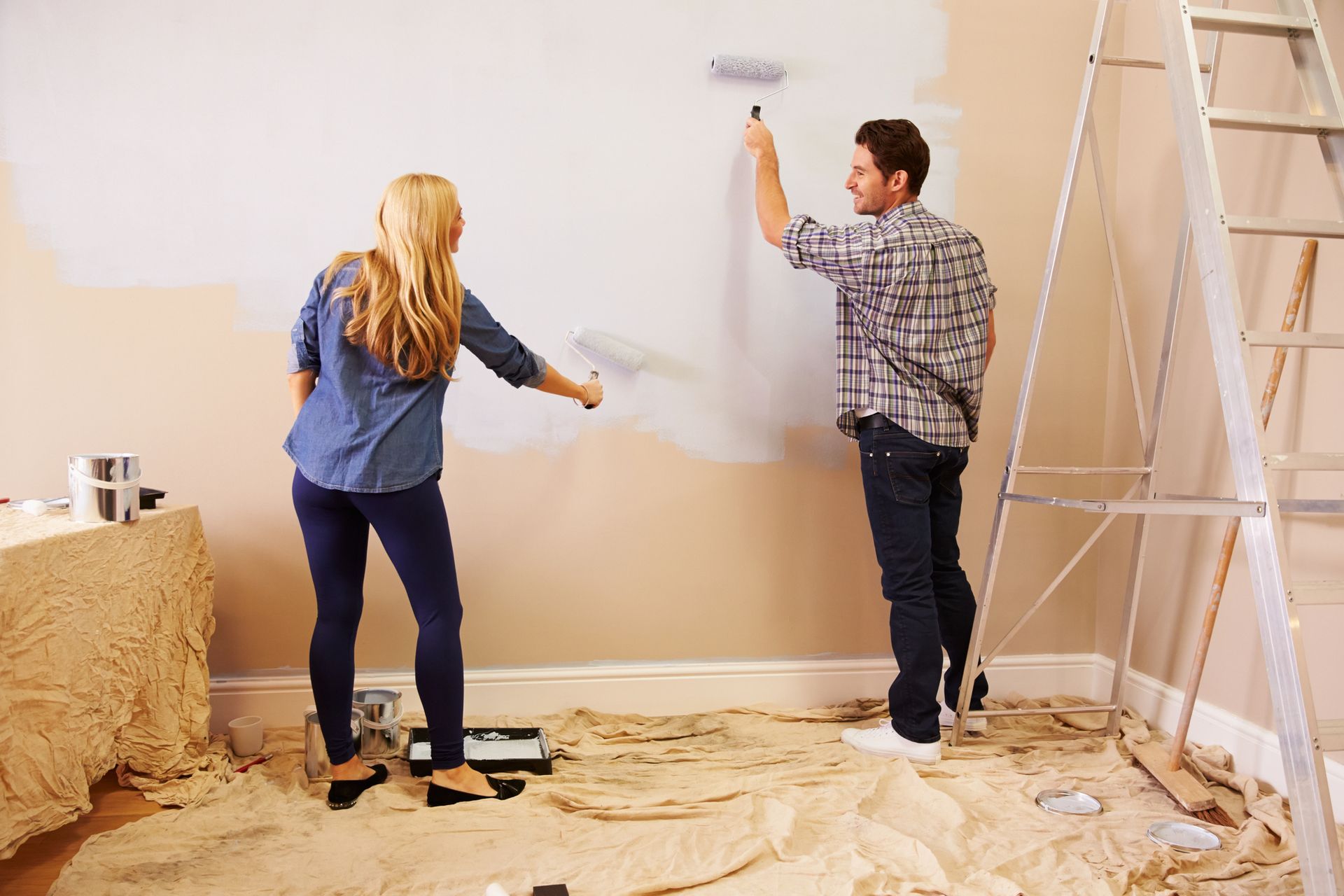 Two people painting a room with beige walls, using rollers beside a ladder and drop cloths.