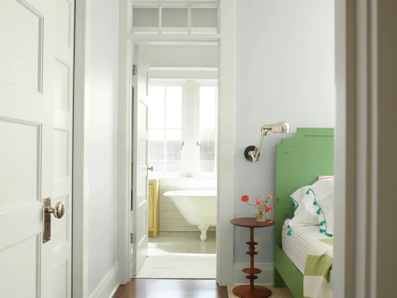 Bright bedroom doorway view into a bathroom, with a green bed, white walls, and a small red stool.