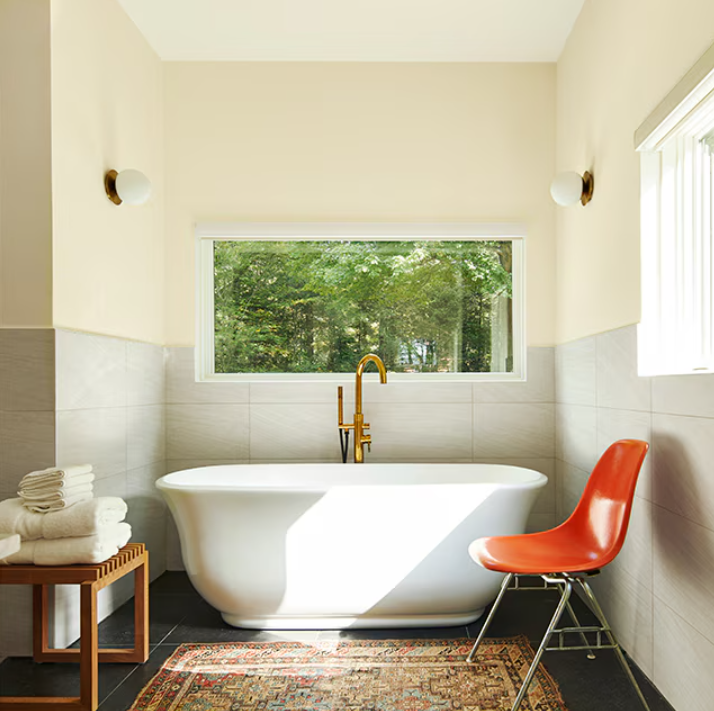 Bright bathroom with a freestanding tub, gold faucet, orange chair, and a large window overlooking greenery