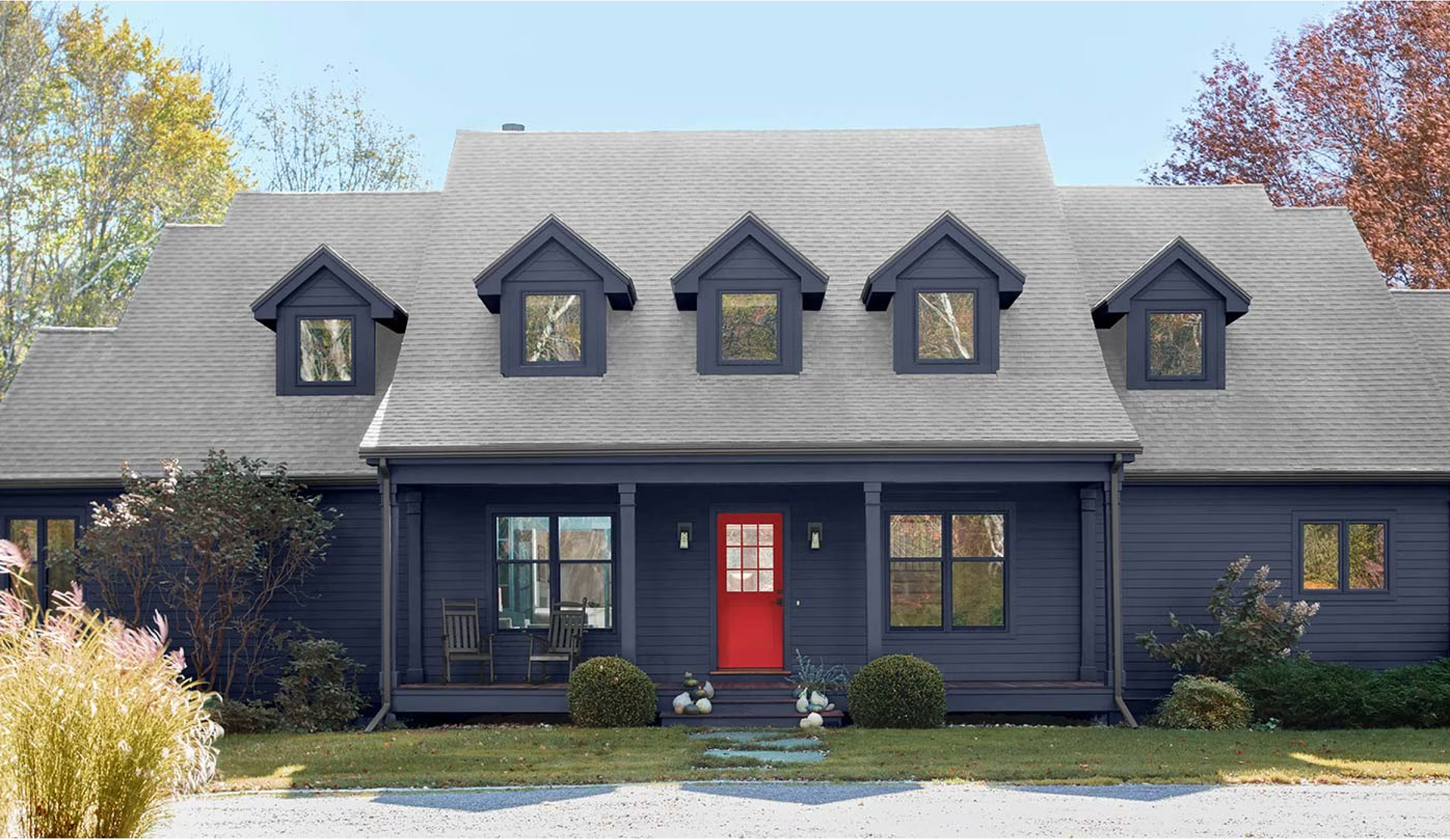 Dark blue house with four dormers and a bright red front door, framed by shrubs and trees.