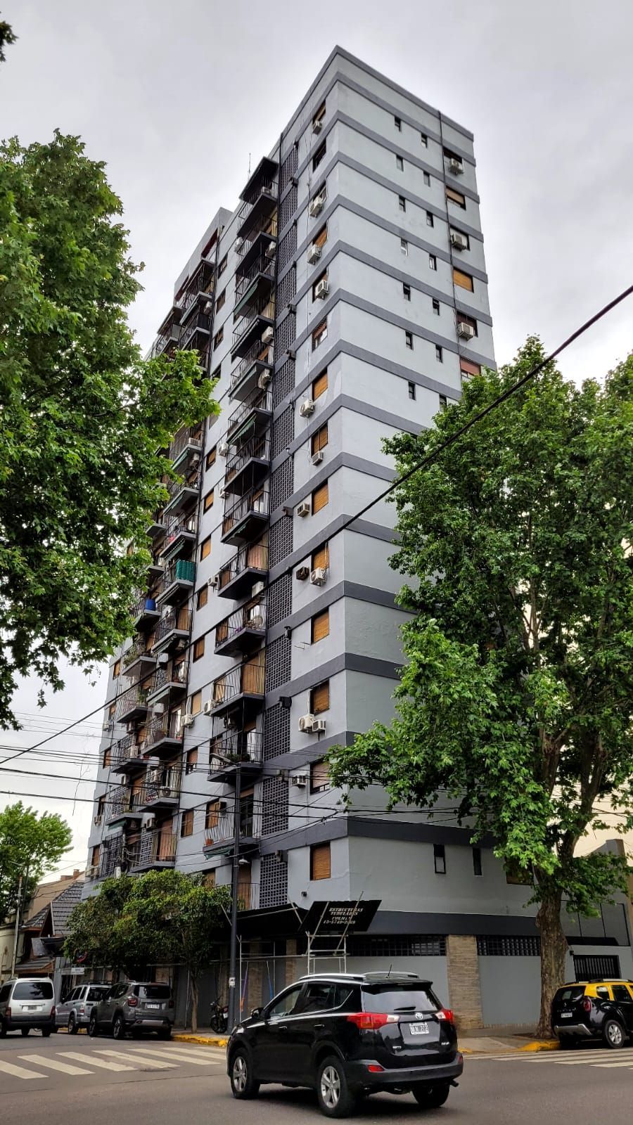 Edificio de apartamentos alto y gris en una calle arbolada de la ciudad con coches aparcados en la planta baja.
