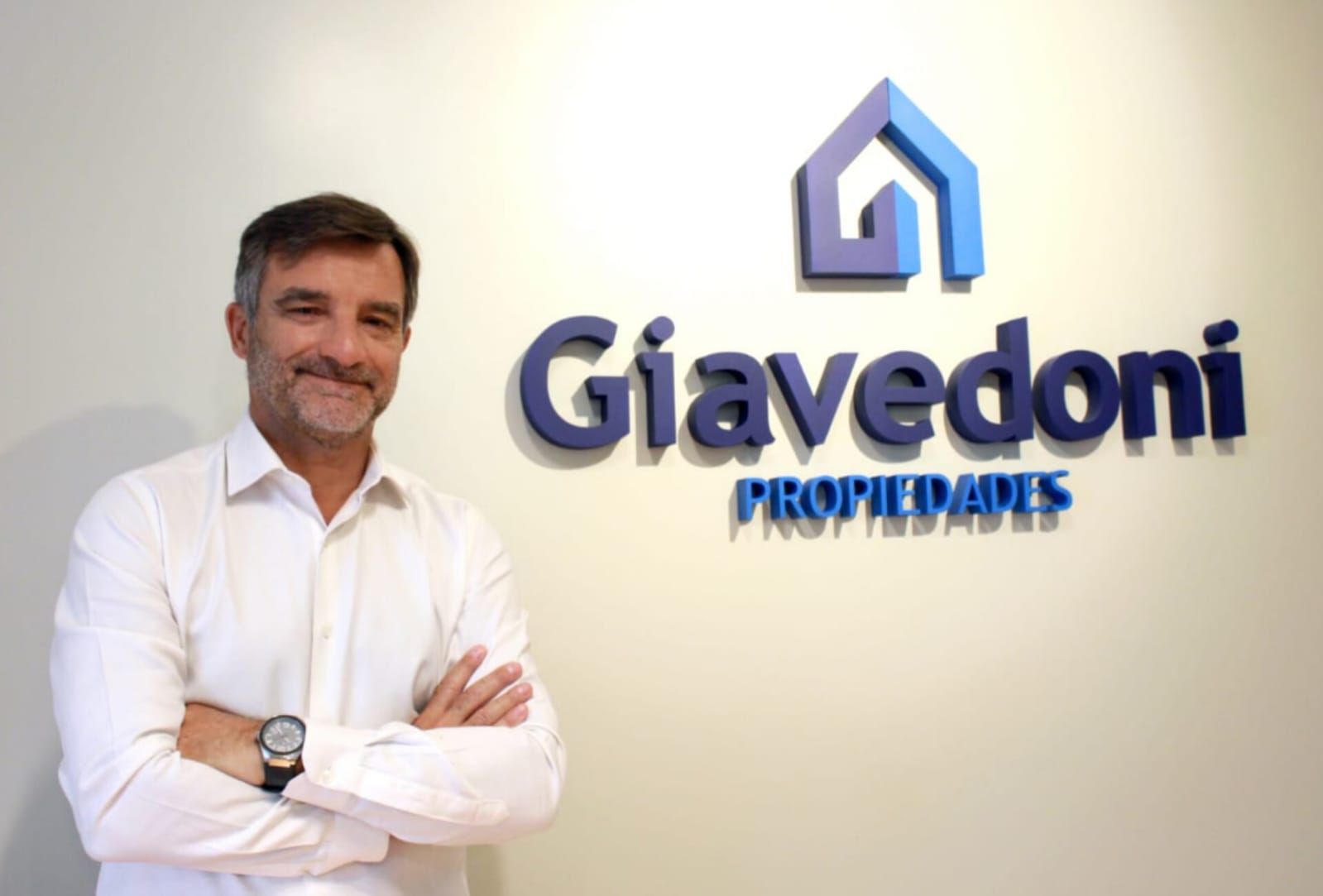 Un hombre con camisa blanca está de pie con los brazos cruzados, junto a un logotipo azul de Giavedoni Propiedades