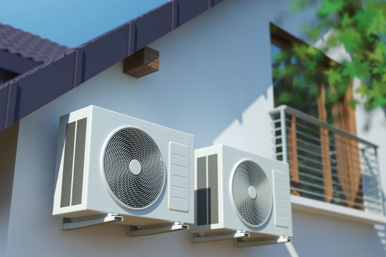 Two white outdoor air conditioner units mounted on a blue house wall near a balcony and window.