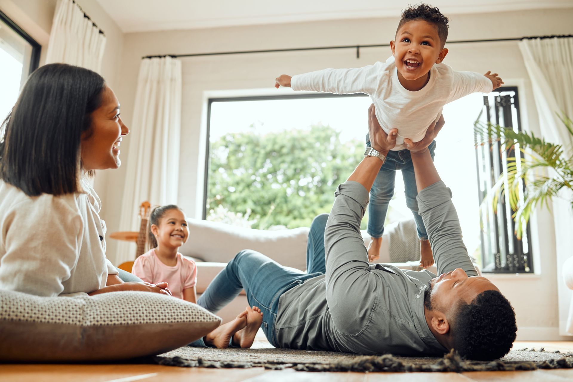 Family playing together in a bright living room, with a child held aloft by an adult lying on the floor.