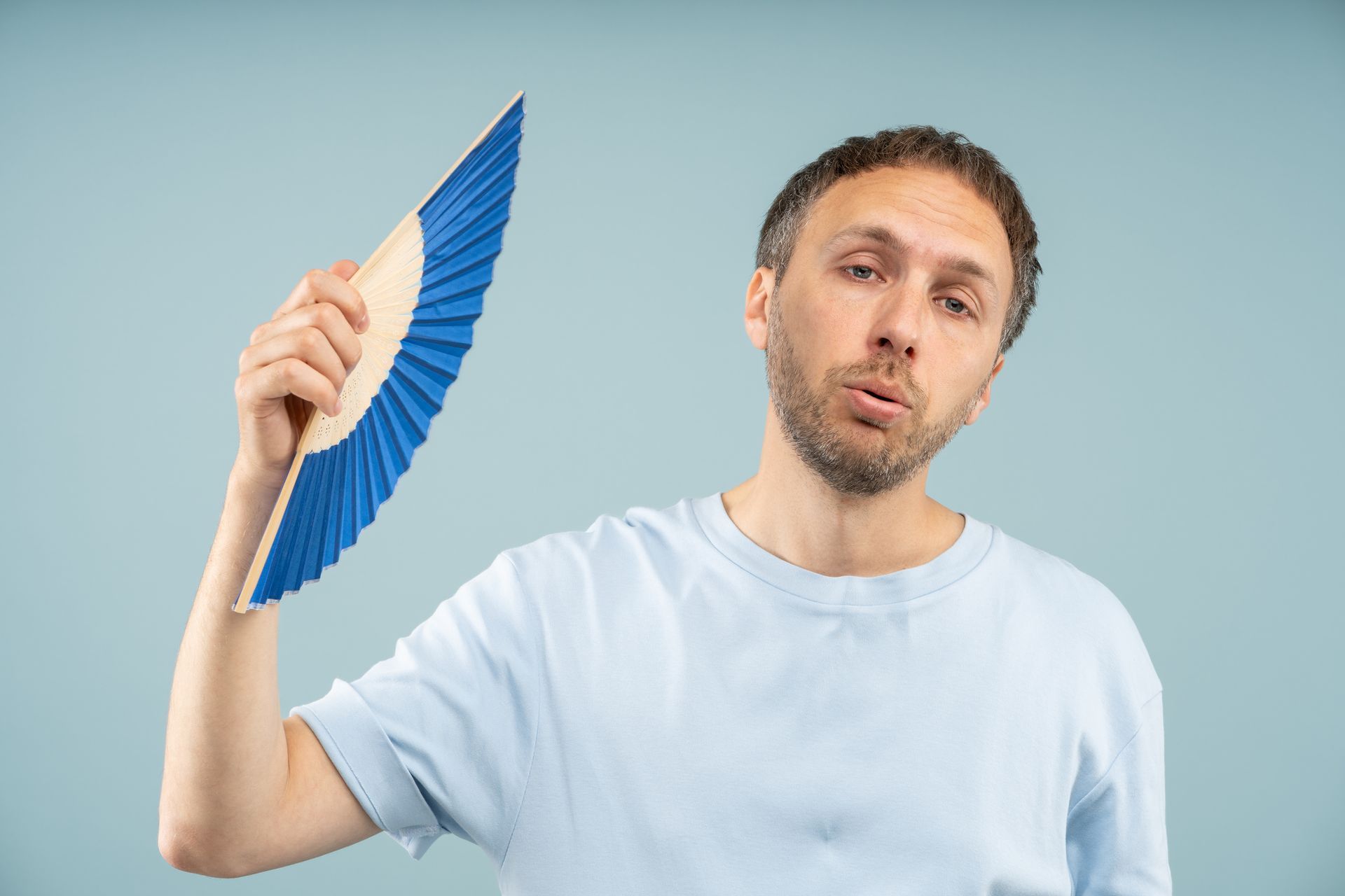 Overheated homeowner using a handheld fan indoors while struggling with an air conditioner.
