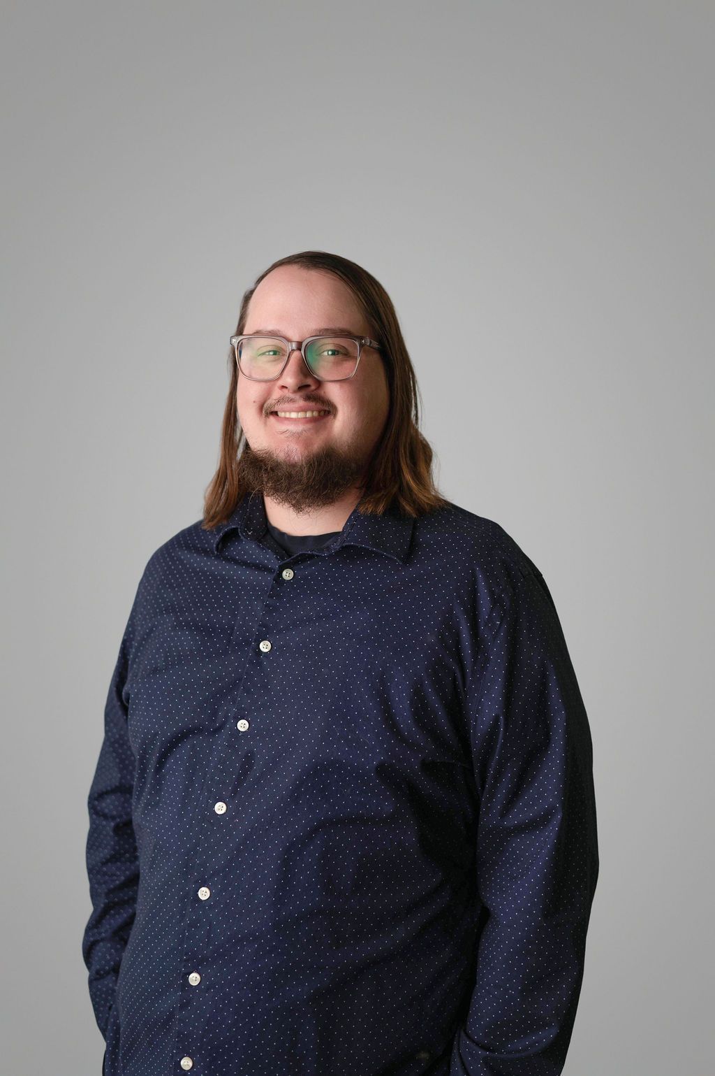 Griffin Robbins with long hair and glasses, wearing a dark blue button-down shirt, smiling in front of a gray backdrop.