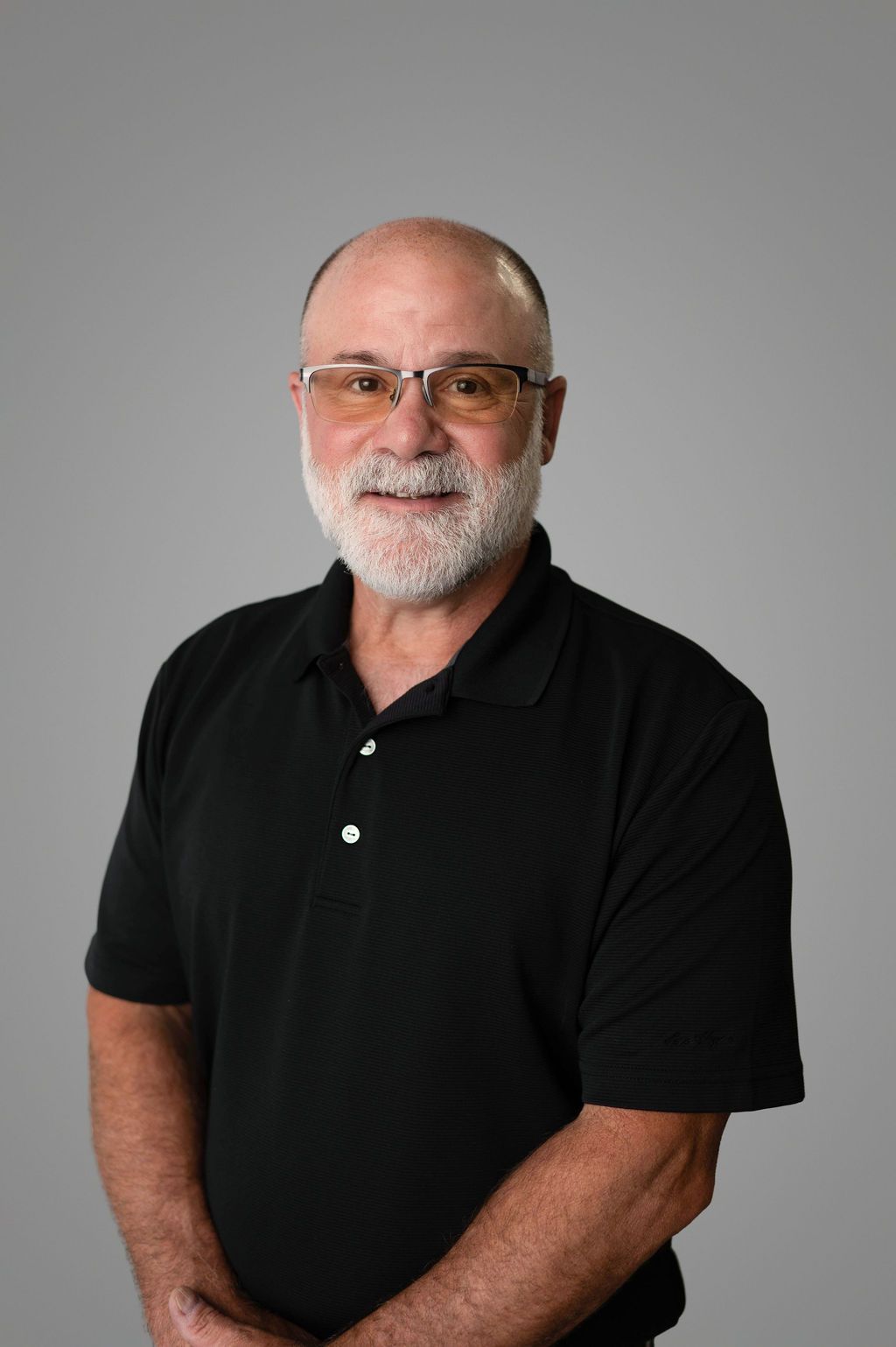John Hubbard in black polo shirt, glasses, and white beard, smiling against a gray backdrop.