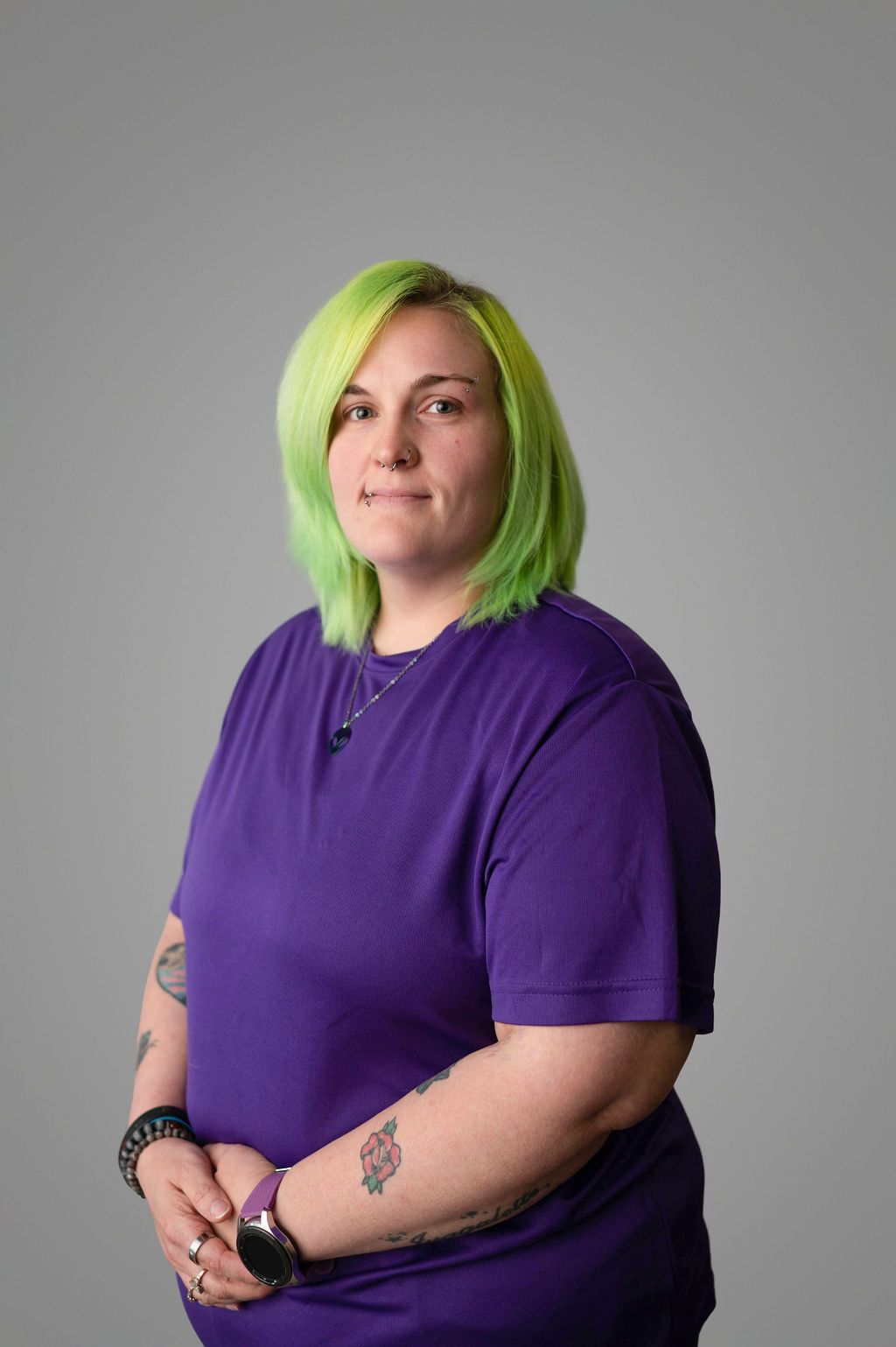 Lexi Dyer with green hair, purple shirt, and tattoos, standing in front of a gray background.