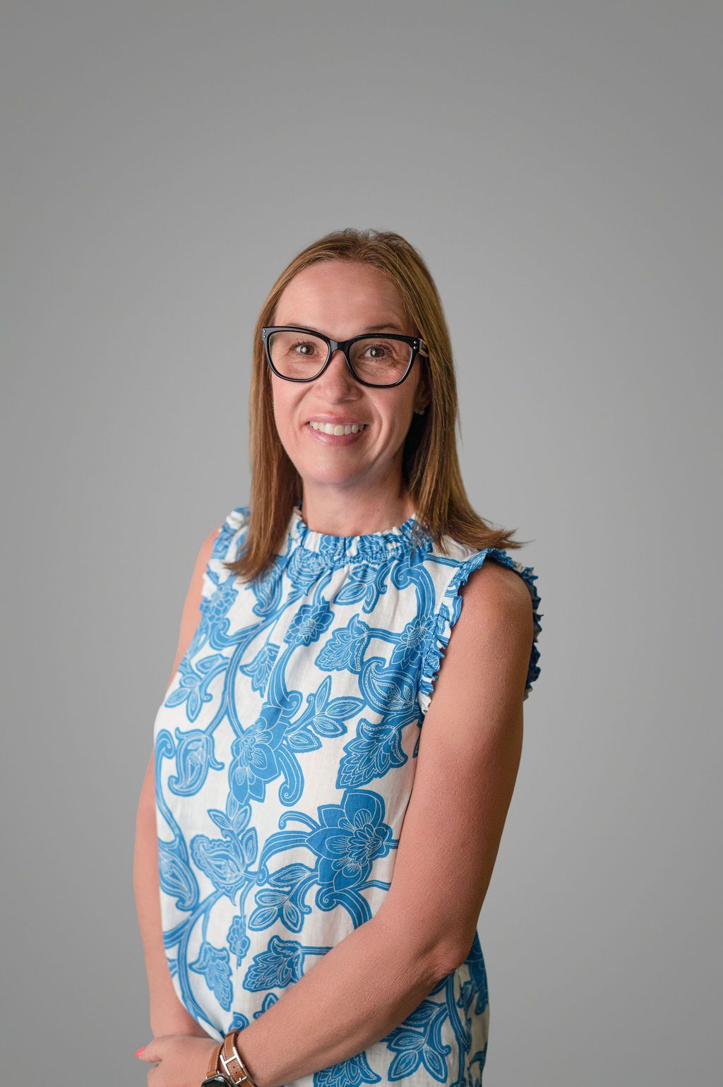 Tara Robbins with glasses wearing a white and blue patterned top, smiling. Against a gray background.