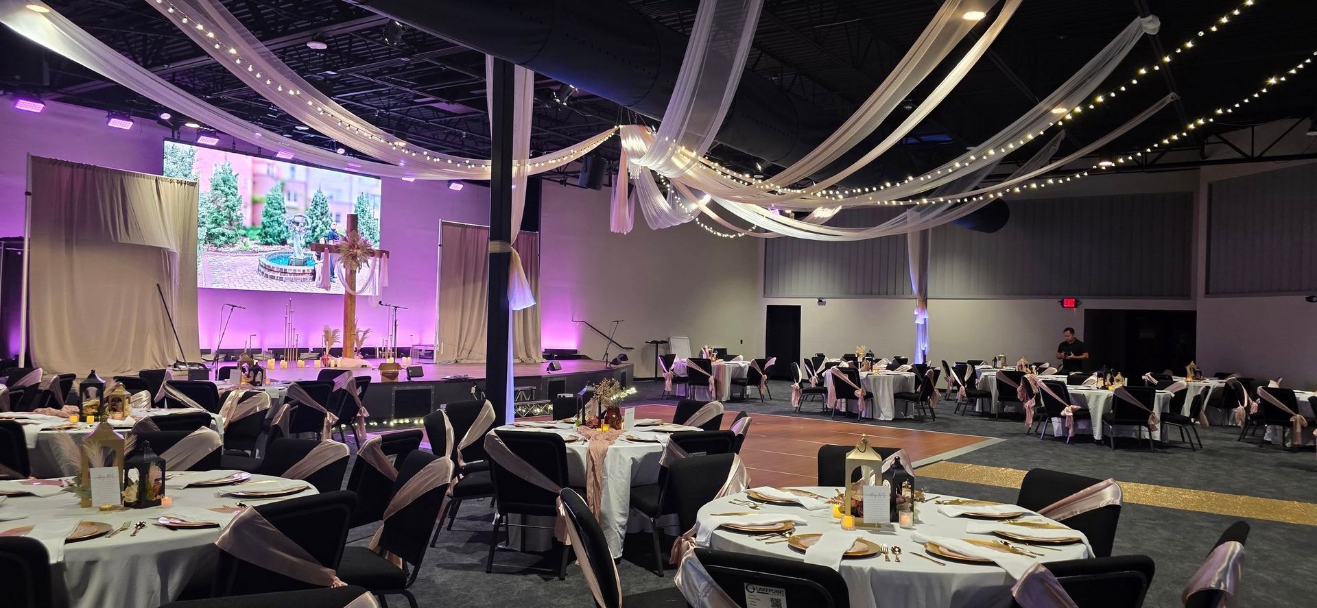 Banquet hall set for an event; tables set, stage with screen and cross, ceiling draped with fabric.
With Love, Wedding management, Milwaukee Wedding Coordinator
