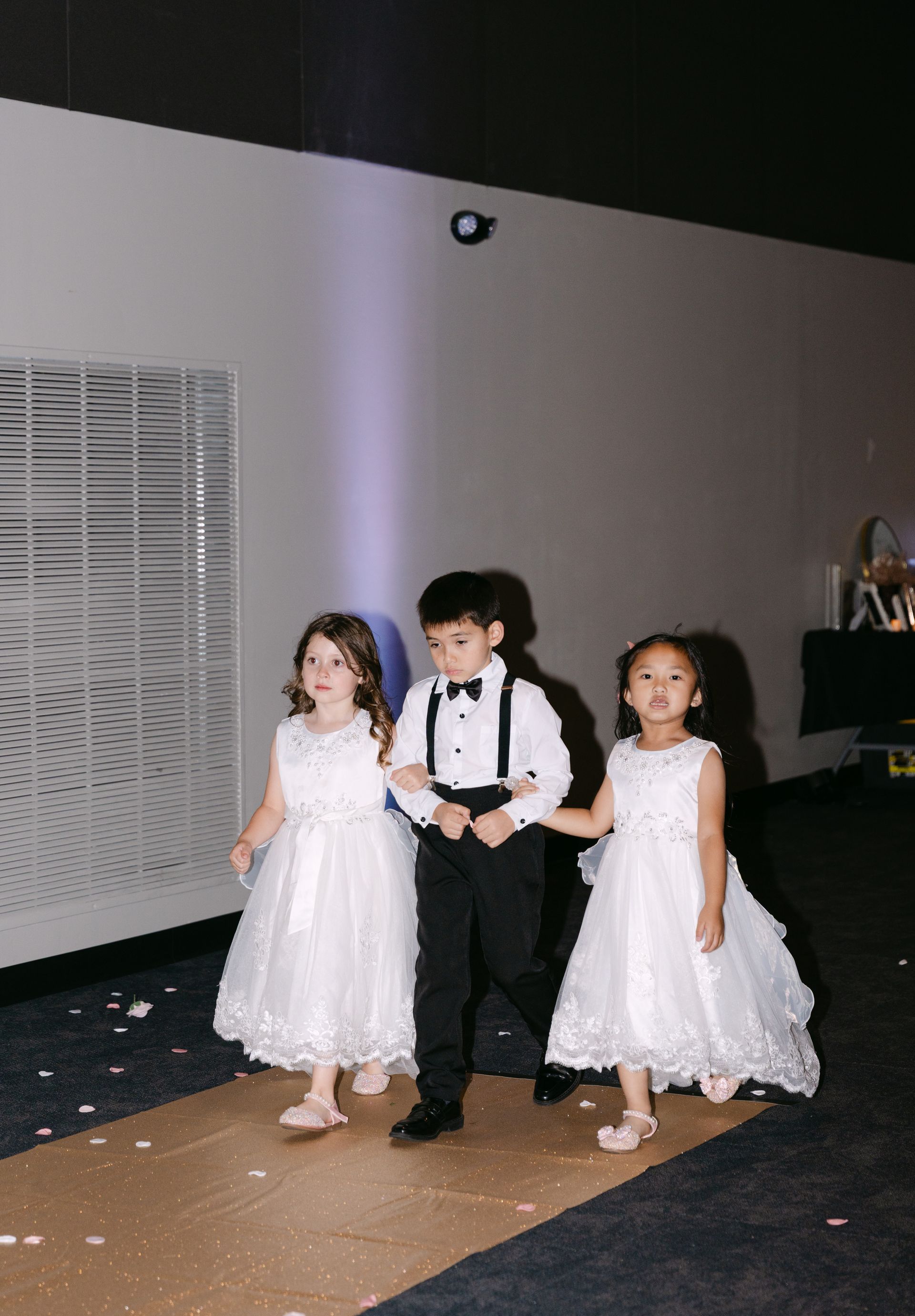 Three children walking down an aisle: boy in tuxedo, two girls in white dresses, carrying flowers.
