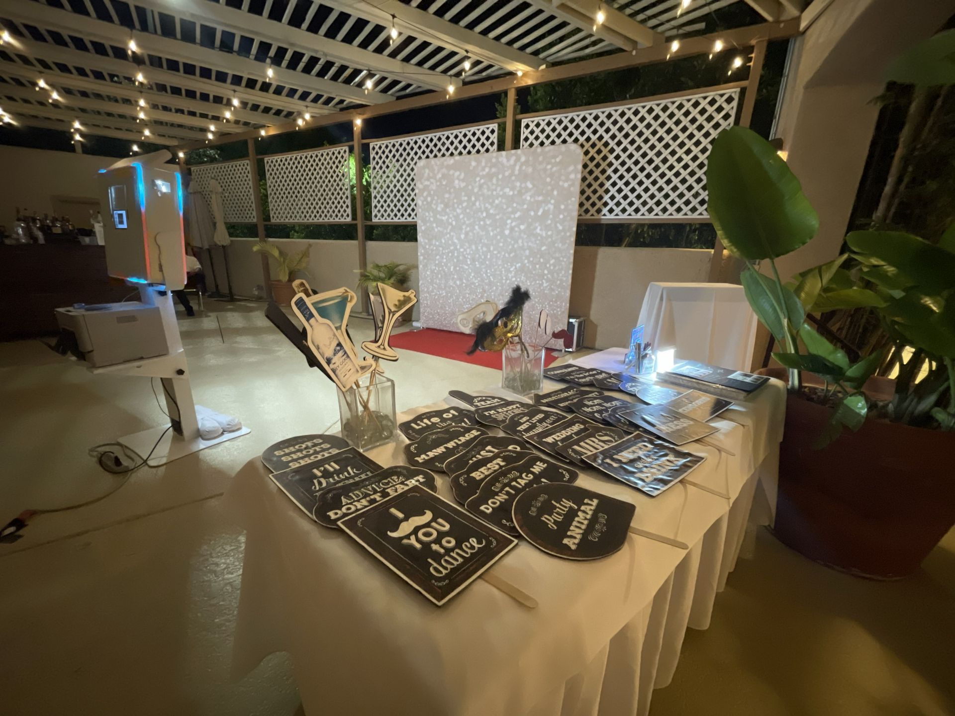 Vendor table with black signs and plants at an indoor market or fair under a covered pavilion.