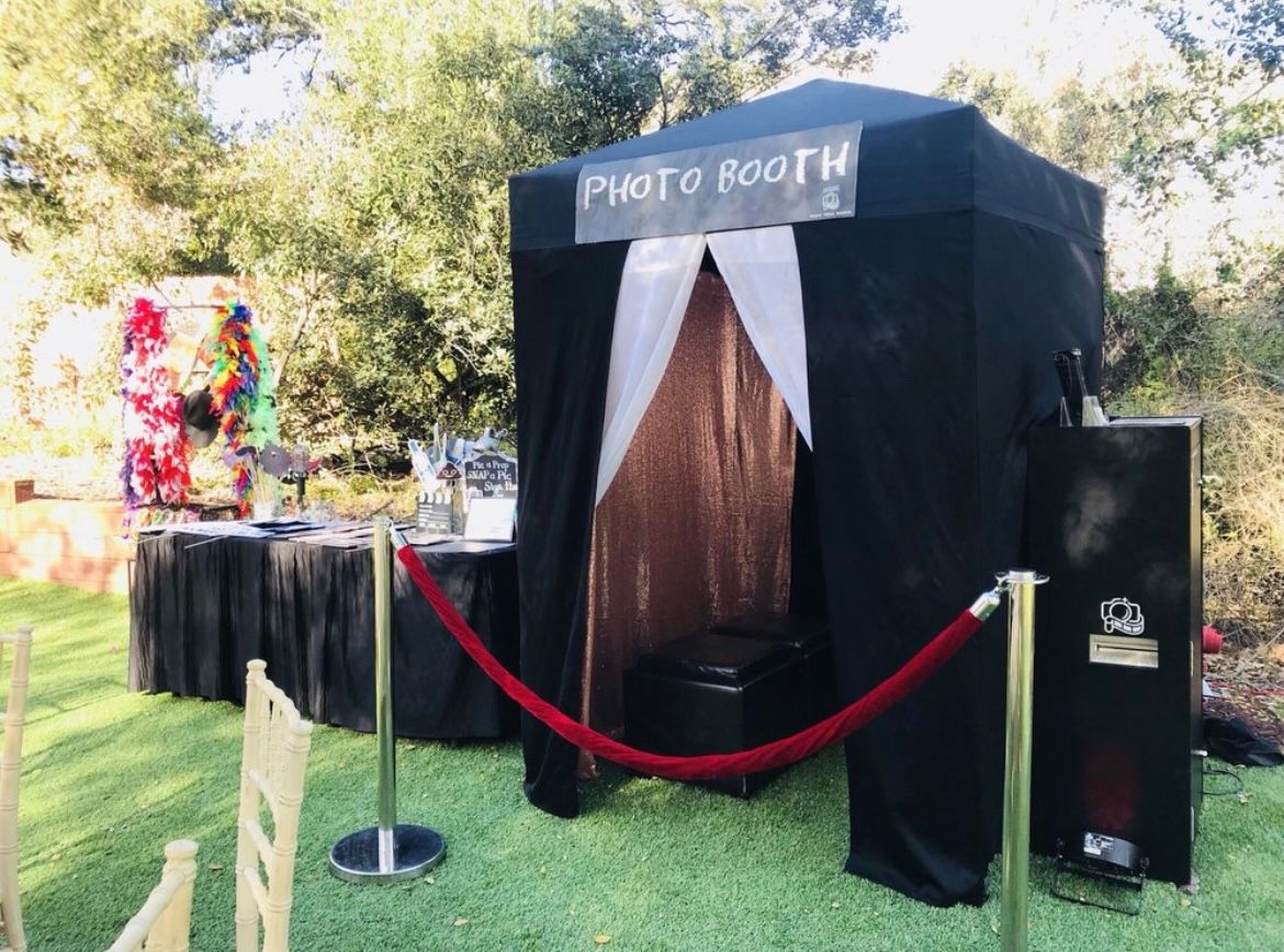 Outdoor photo booth with black drapes, white curtains, and red rope barriers on a grassy lawn.