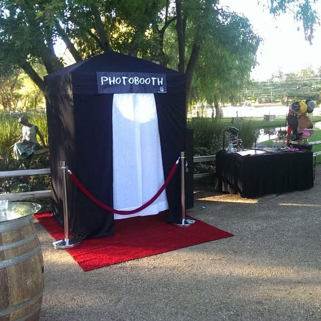 Outdoor photo booth with black drapes, red carpet, and a table beside it in a park.