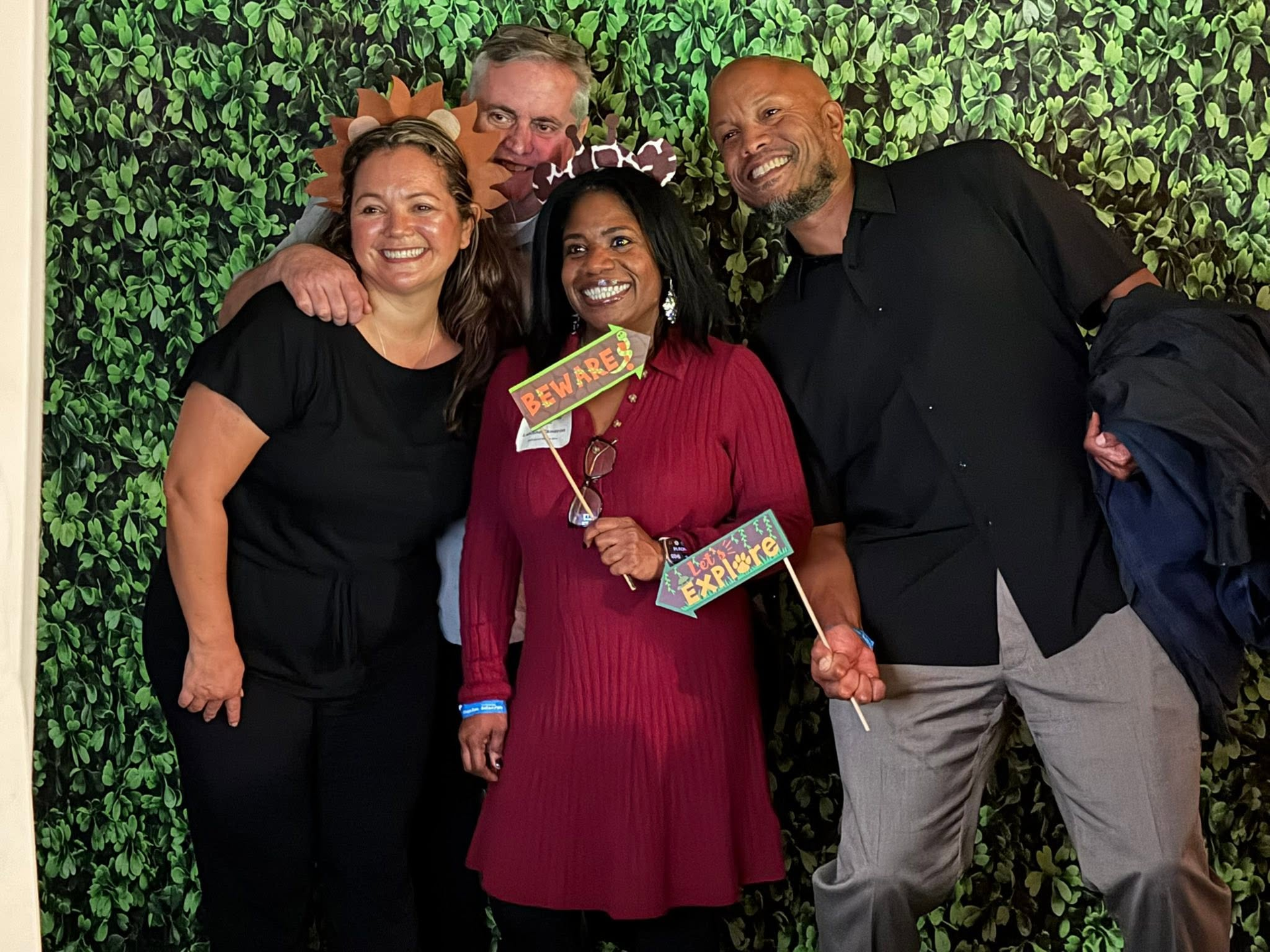 Four smiling people posing together in front of a leafy green backdrop, holding colorful signs.