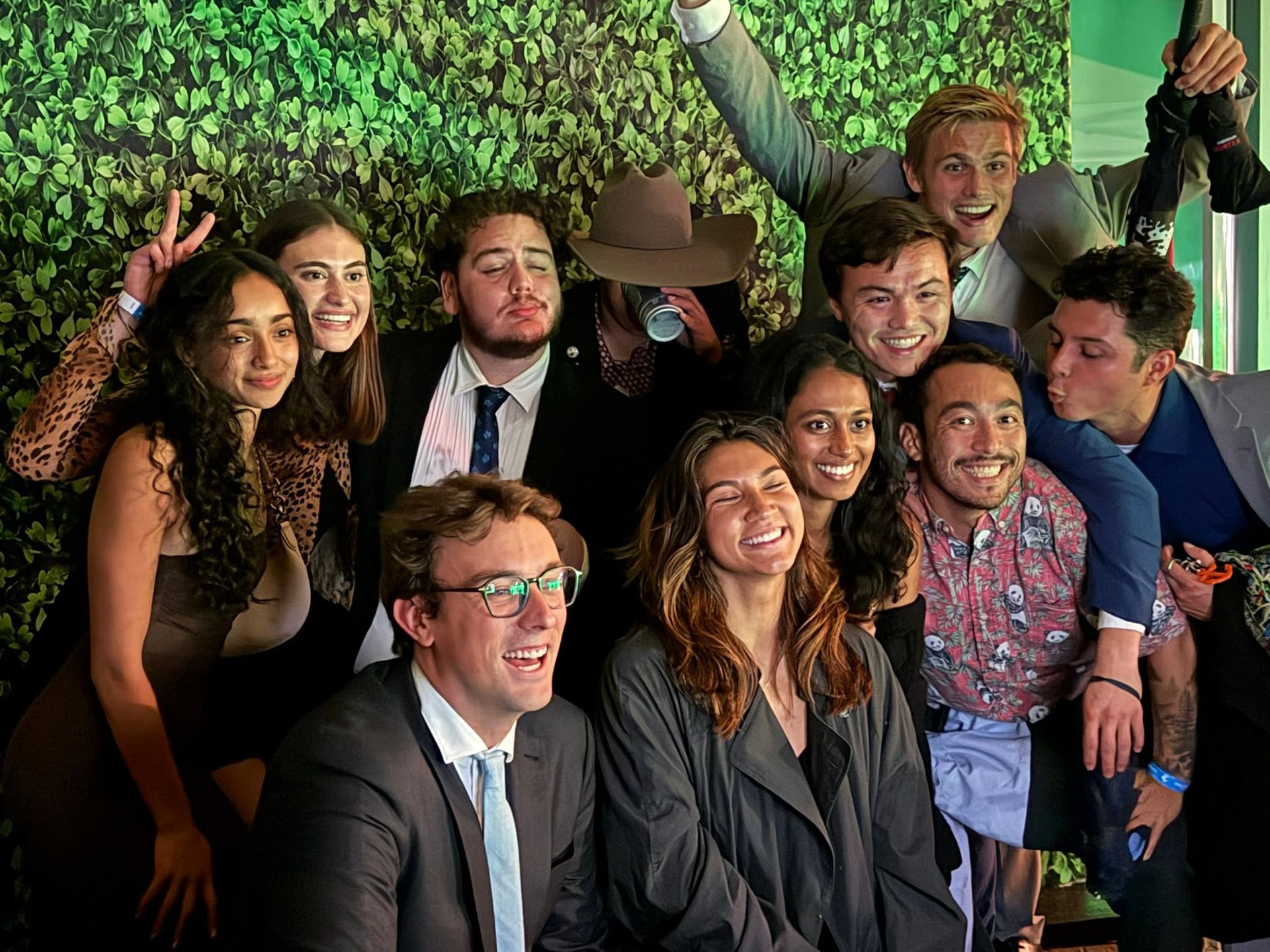 Group of people smiling and posing together in front of a green plant wall at an indoor event