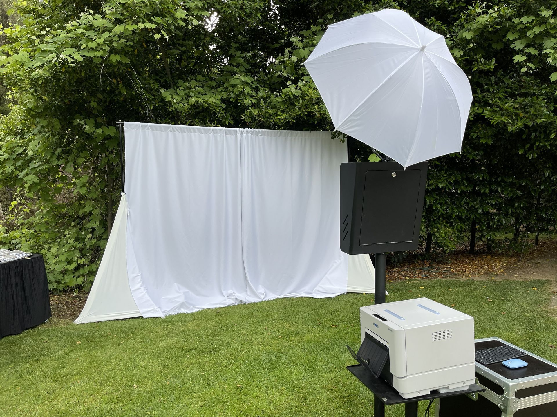 Outdoor photo booth setup with white draped backdrop, umbrella lights, and grassy lawn behind trees