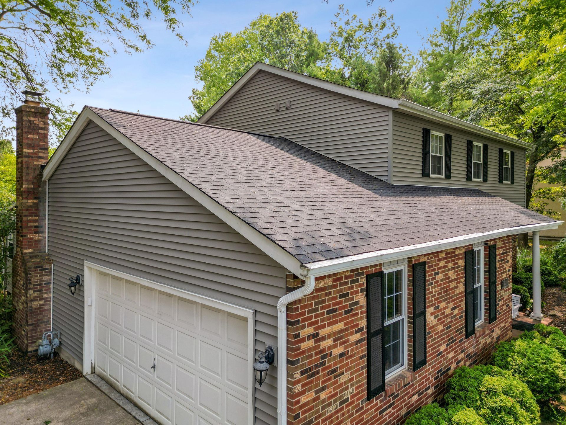 Suburban two-story brick and siding house with white garage door and gray shingled roof under trees