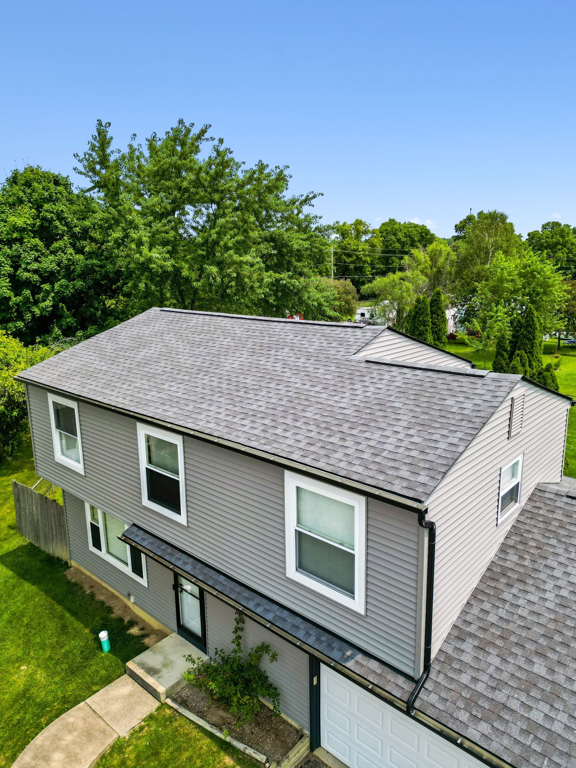 Gray two-story house with black shutters and a sloped roof, viewed from above amid green trees