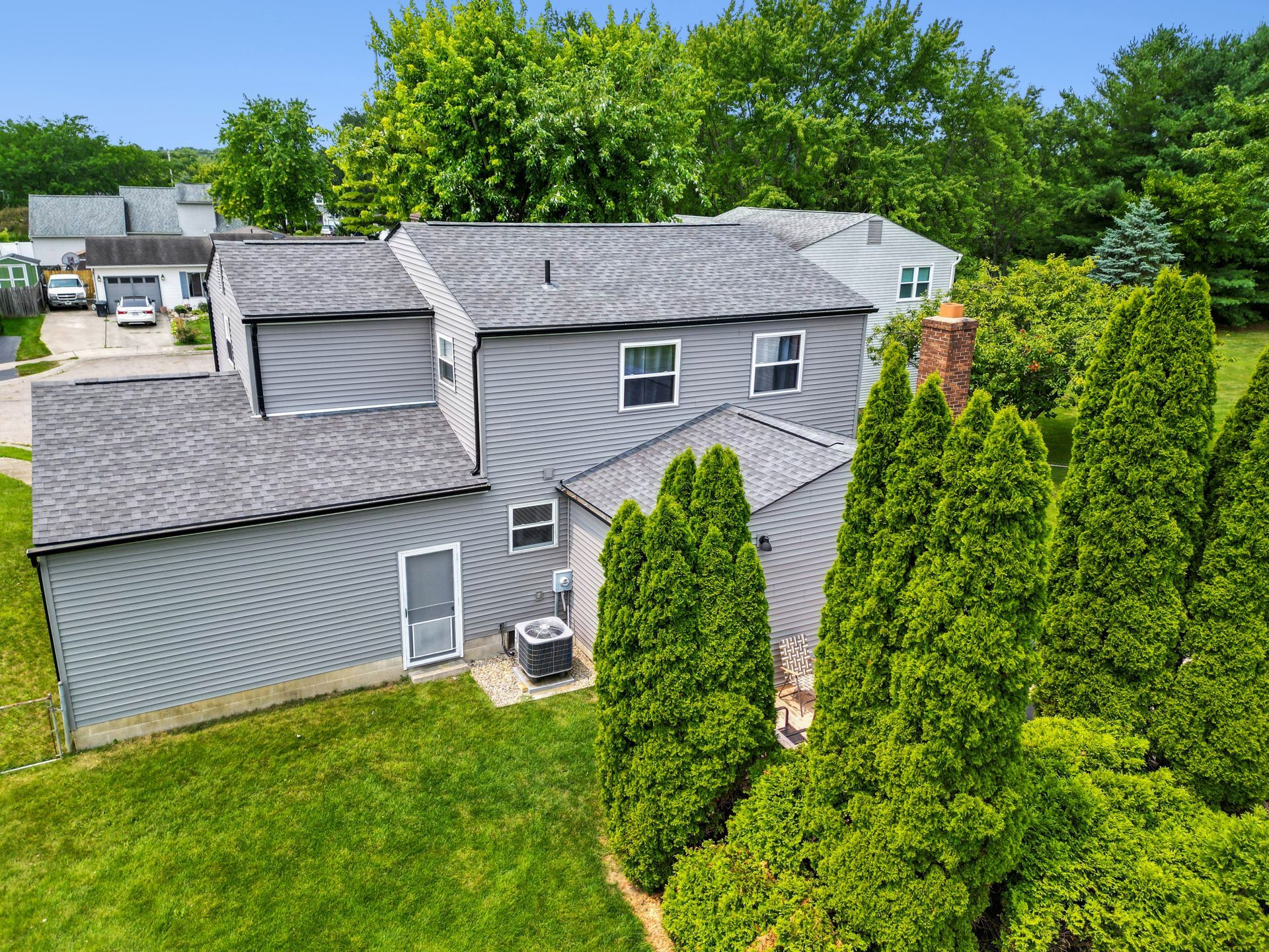 Gray two-story house with a roof deck, green lawn, and tall trees in the foreground