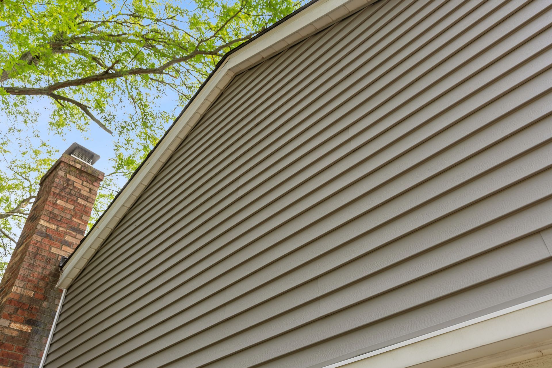 House exterior with beige siding, brick chimney, and green tree branches overhead