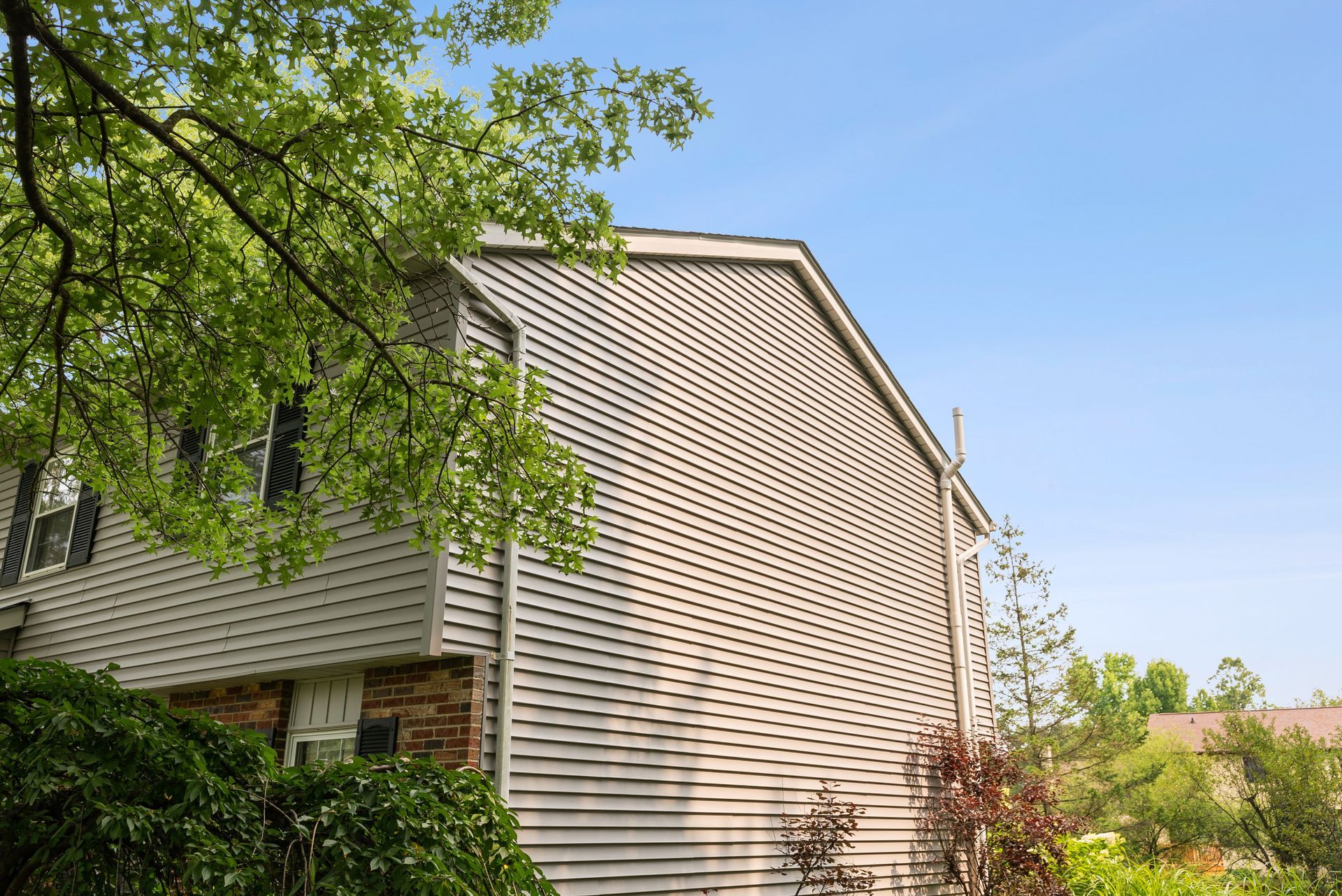 Side of a beige house with green trees and shrubs under a blue sky