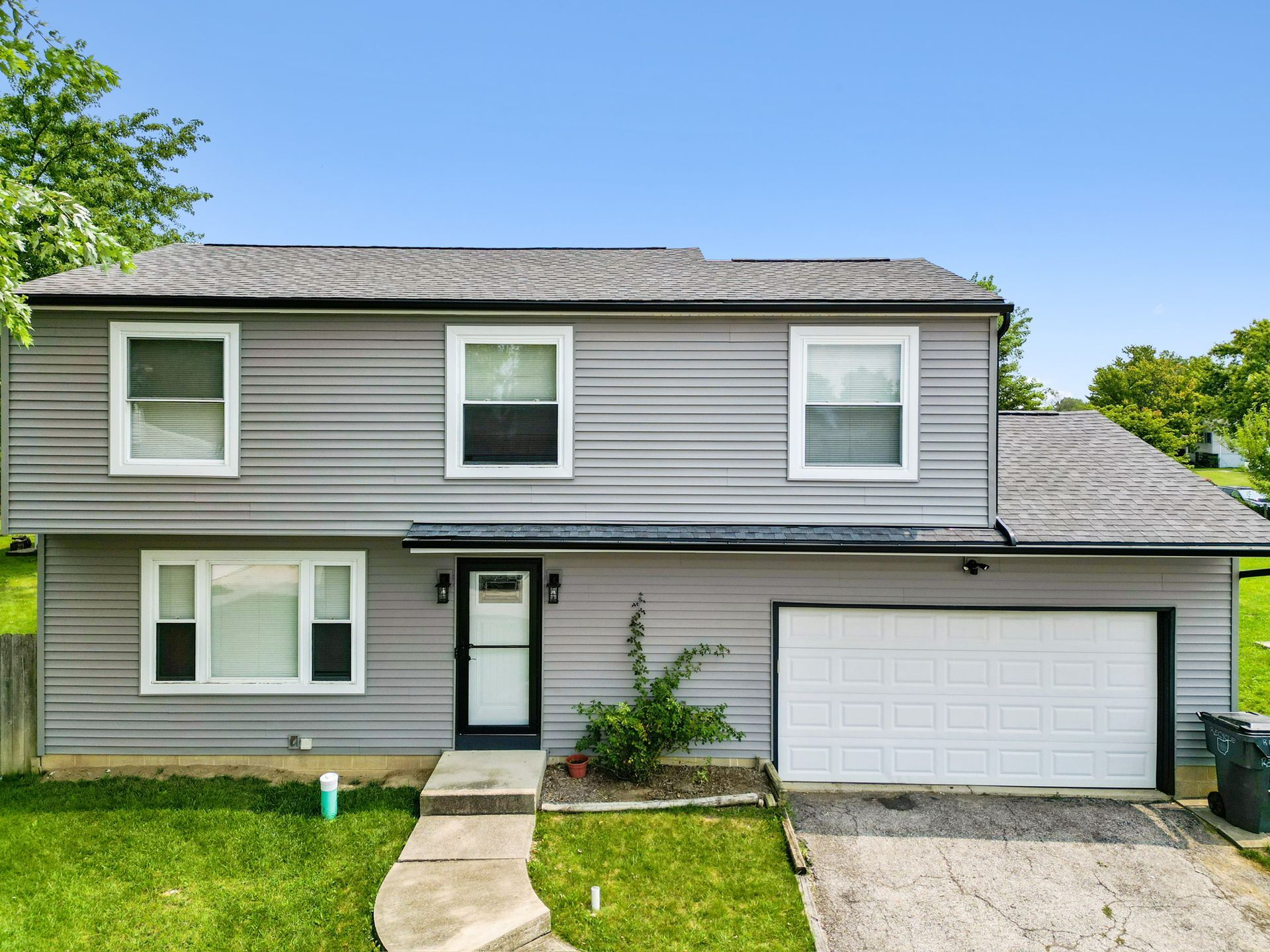 A two-story house with grey siding, a white front door, a white garage door, and a grassy front lawn under a clear sky.