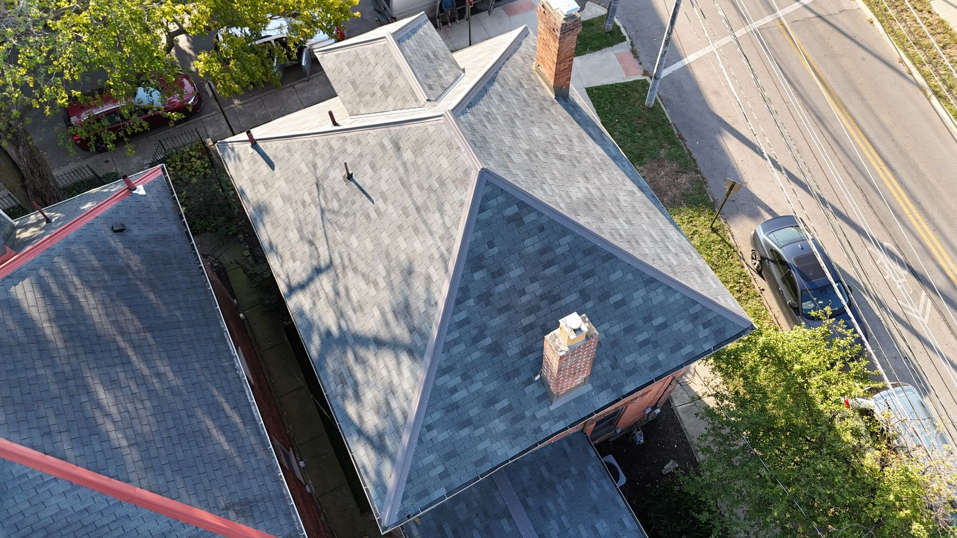 Aerial view of gray shingled roofs on residential houses, with chimneys and a street with a car visible nearby.