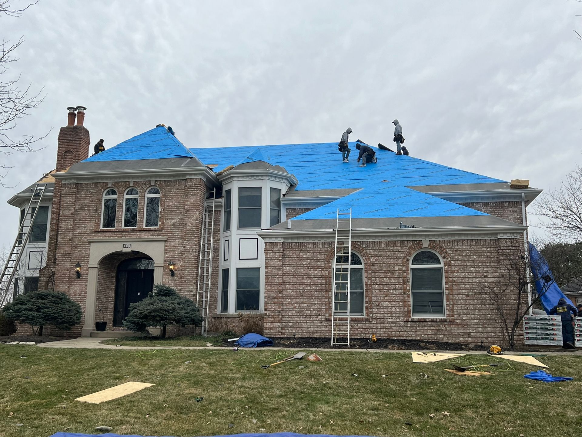 Workers on the roof of a large brick house covered in blue protective tarp, with ladders leaned against the exterior.