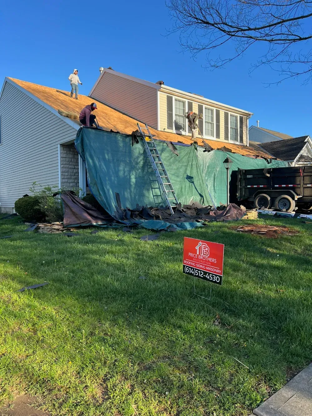 Construction workers replacing the roof of a house, with a green tarp covering the siding and a dumpster nearby.